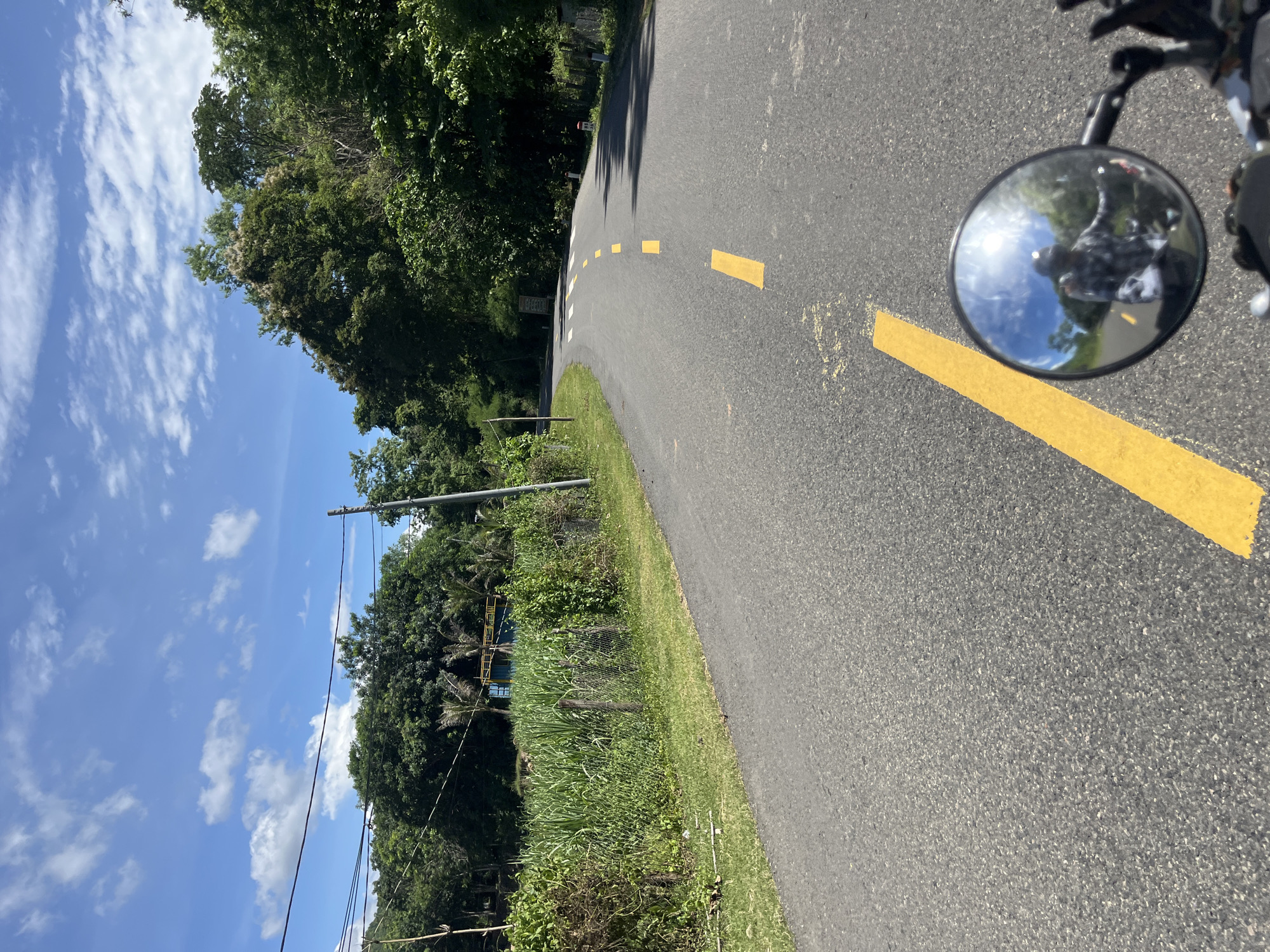 The image captures a view from a motorcycle traveling on a paved road in Ham Thuan Bac, Vietnam. The road, marked with yellow dashed lines, curves to the left, disappearing into a lush, green landscape. The sky above is a vibrant blue, dotted with scattered white clouds. On the left side of the road, a grassy embankment leads to a field of tall, green crops, possibly sugarcane, bordered by a wire fence. Beyond the field, trees and a small structure are visible. The right side of the road is lined with dense, mature trees. In the foreground, a circular motorcycle mirror reflects the rider, wearing a helmet and a checkered shirt, and the sunny sky. The overall scene depicts a scenic rural road on a clear day.