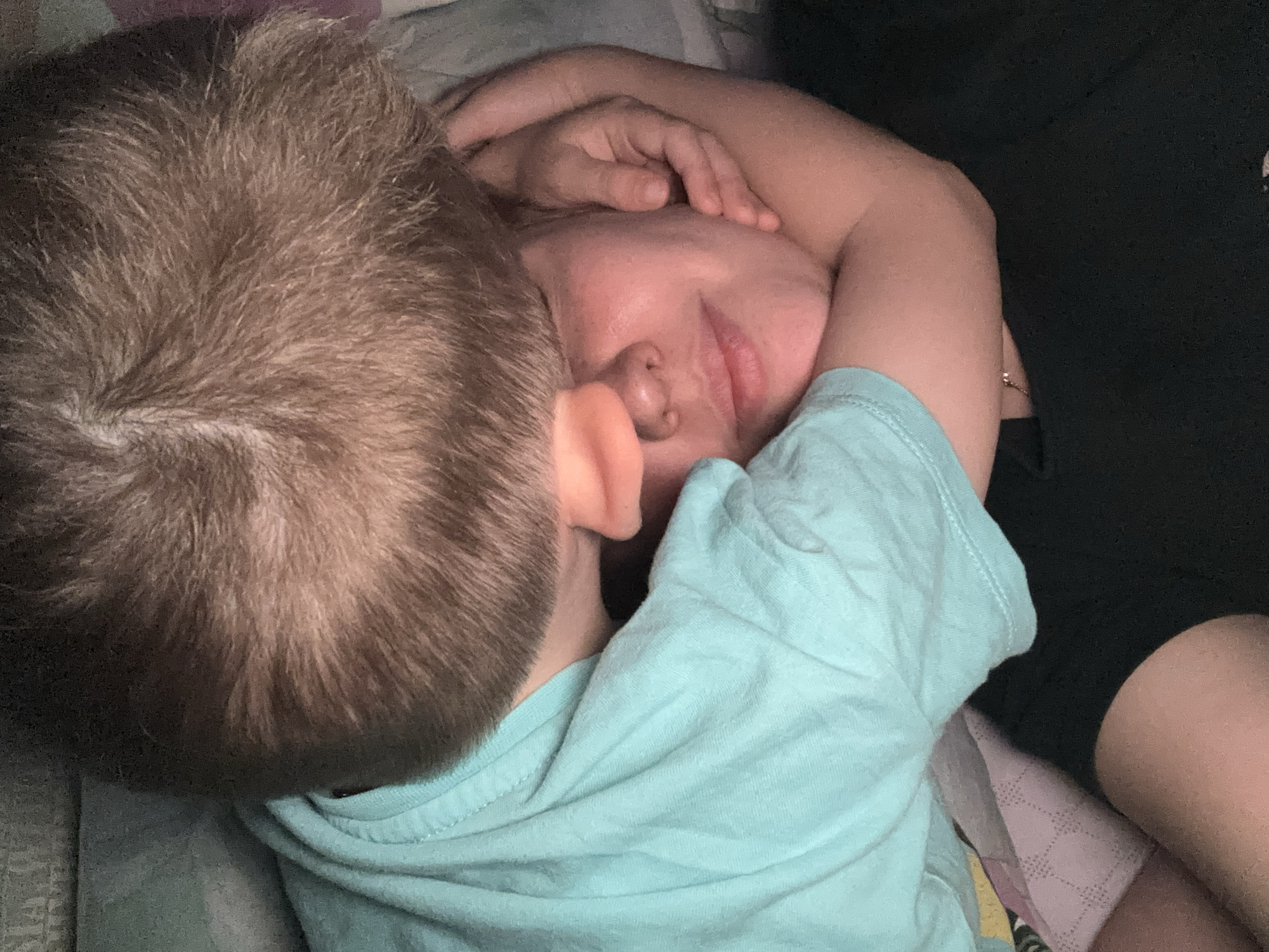 A child with short brown hair and wearing a light blue shirt is lying with their head on an adult's face. The adult is lying on their back with their eyes closed and one arm around the child. The adult has fair skin and visible lips and nose. They are lying on a bed with patterned sheets. Part of another person wearing black pants or shorts is also visible in the upper right corner of the image. The location in Rusanovo, Russia is not determinable from the image.