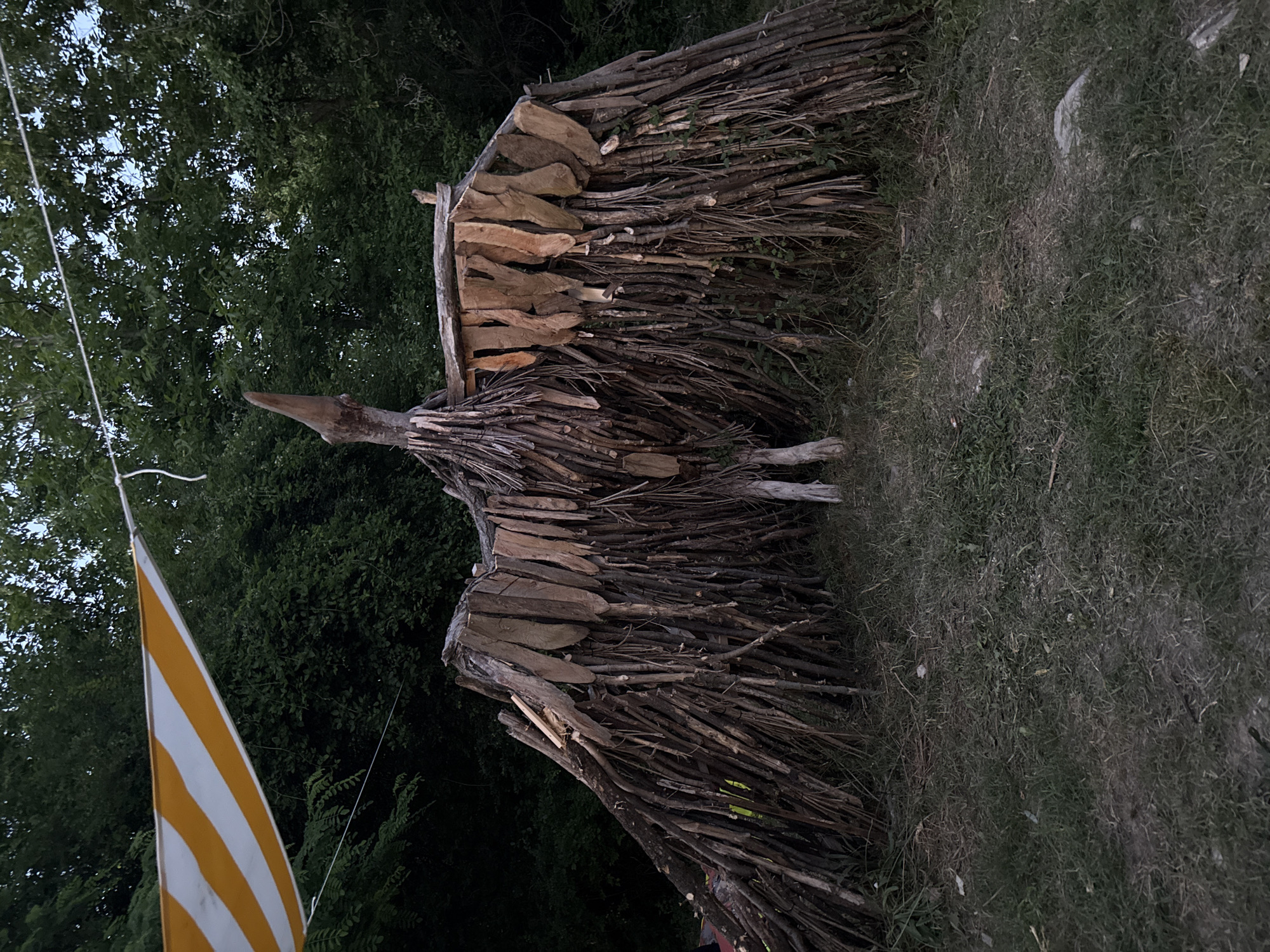 A large, avian-shaped sculpture made of natural materials stands in an outdoor setting. The sculpture is constructed from numerous twigs and branches, forming a body, wings, and a head with a beak. Flat wooden pieces are attached to the top of the wings, resembling feathers. The sculpture is supported by two wooden legs. In the background, a dense forest of green trees is visible. A yellow and white striped tarp is suspended by a rope above and to the left of the sculpture, casting a shadow. The ground is covered in grass and dirt. The scene appears to be an artistic installation or a temporary structure in a natural environment. No specific location in Stara Zagora, Bulgaria, can be identified from the image alone.