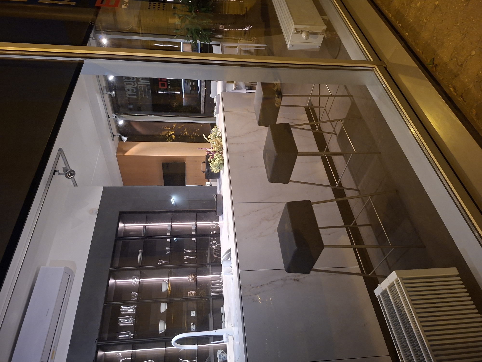 This is a photograph of a kitchen showroom display, viewed from the exterior through a large glass window. The showroom appears to be a modern, upscale establishment.
In the foreground, three bar stools with dark, square seats and thin metal legs are arranged in front of a sleek, marble-patterned kitchen island. The island features a white, contemporary faucet. Behind the island, a wall unit with glass-fronted cabinets is illuminated from within, displaying various glassware and dishes. The cabinets are recessed and have a dark frame. Above the cabinets, a white air conditioning unit is mounted on a dark grey wall.
Reflected in the glass window, a street scene is visible, including a building with signage that reads "neog In". The exterior appears to be at night, with streetlights casting yellow illumination. A white radiator is positioned on the right side of the frame, near the window. The overall impression is one of a well-lit, organized interior space contrasted with the darker, illuminated exterior.
While the image is presented as a kitchen showroom, and the context provided suggests it might be in Tallinn, Estonia, there are no explicit visual cues within the photograph itself (such as street signs, landmarks, or language on the signage) that definitively confirm the location as Tallinn, Estonia. The signage "neog In" is the only text visible and does not provide a clear geographical identifier.