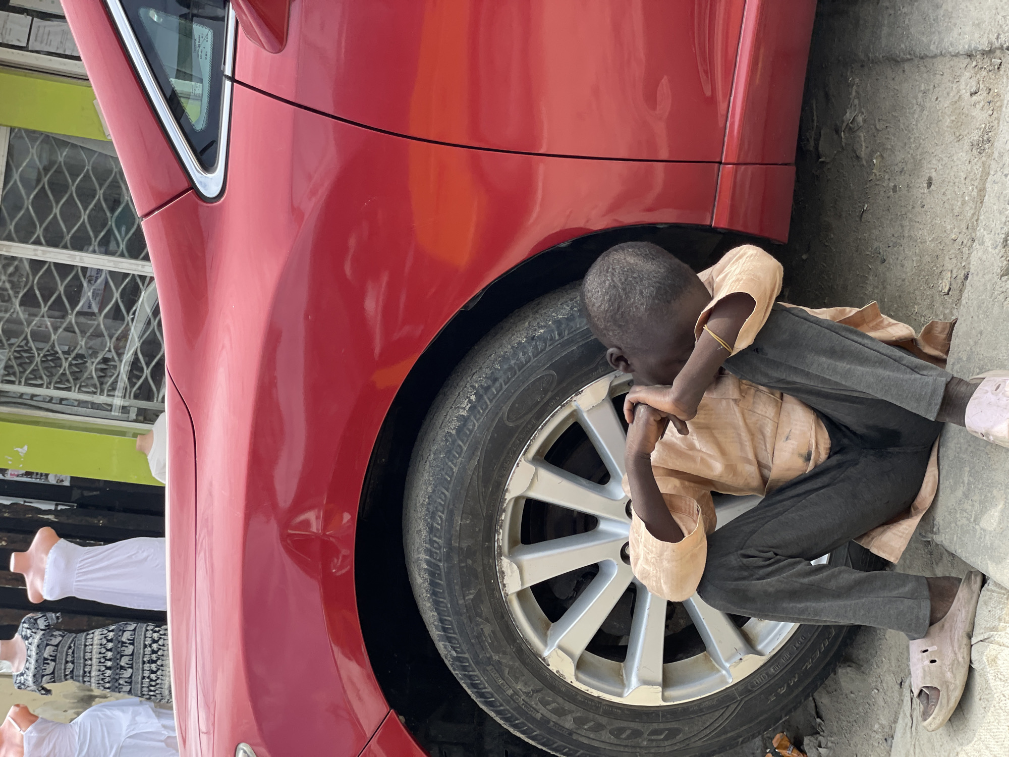 A young boy is seen sitting on the ground next to a red car. He is wearing a light brown shirt and dark grey pants, with a yellow band on his left wrist. His head is bowed and he appears to be resting, with his arms wrapped around his knees. The boy is wearing light pink sandals. The car has a visible tire and wheel. In the background, to the left of the car, there are mannequins dressed in white and patterned clothing, suggesting a shop or market. The ground is rough concrete. The scene appears to be outdoors, with what looks like natural light, possibly indicating daytime. The location is identified as Lagos, Nigeria.