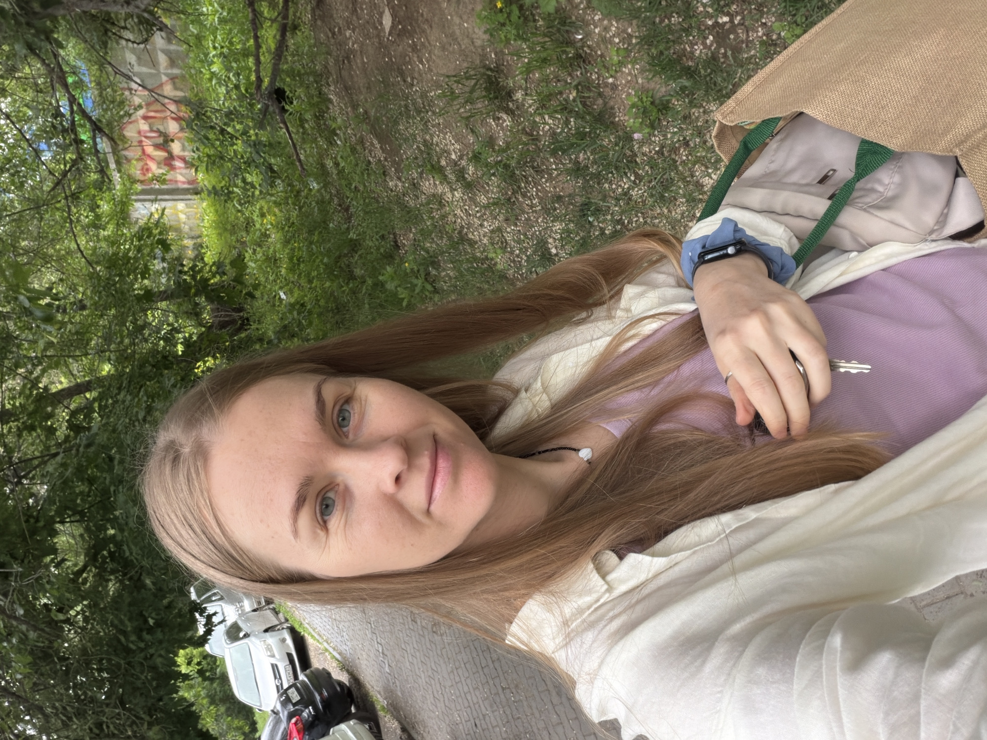 A woman with long blonde hair, wearing a light-colored shirt over a purple top and a necklace, is taking a selfie outdoors. She is holding a set of keys in her right hand. To her left, a paved sidewalk and parked cars are visible. Behind her, trees and greenery form the background, with a concrete wall with graffiti visible in the distance. The image appears to be taken in Khabarovsk, Russia, possibly near a residential area or a park given the presence of cars and the natural surroundings.