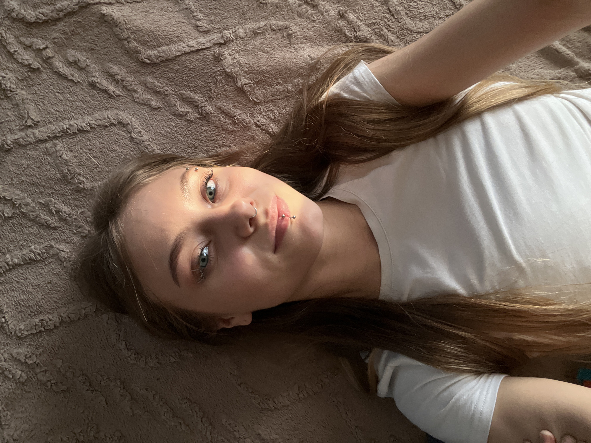 A young woman with long brown hair is lying on a brown textured surface, possibly a bed or blanket. She is wearing a white t-shirt. She has piercings on her eyebrow, nose, and lip. The image is taken from a high angle, looking down at her. The lighting suggests a natural light source from the left.
There is no indication of the city of Фоки, Russia in this image.