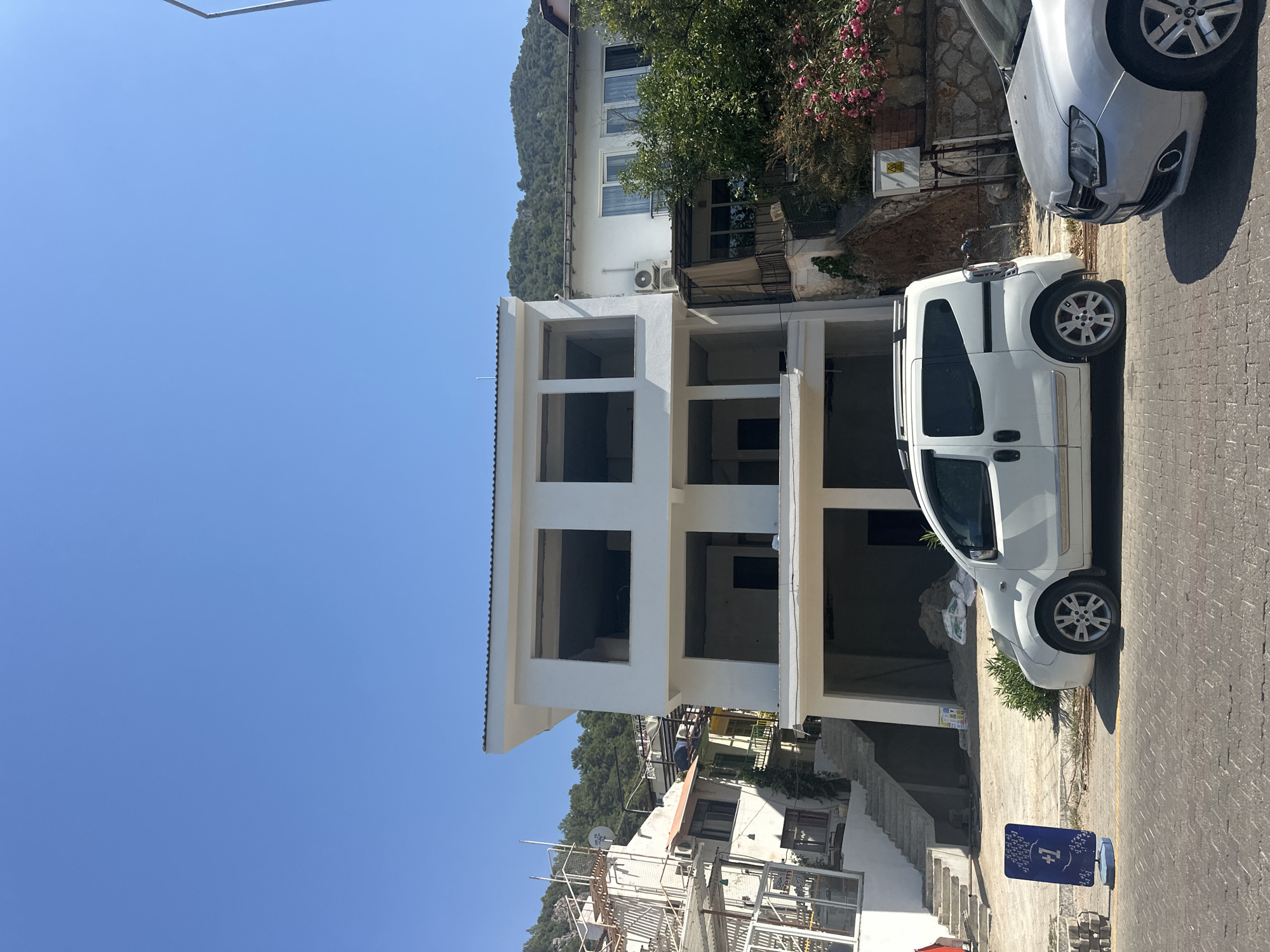 The image shows a street scene in Fethiye, Turkey, on a clear, sunny day. In the foreground, a white minivan is parked on a cobbled street. To the right of the minivan, the front of a silver sedan is visible. Behind the vehicles, a multi-story building is under construction, with open window frames and bare concrete structures. To the left of the construction site, another building with balconies and white walls is visible, with steps leading up to it. In the background, a lush green hillside with trees rises under a bright blue sky. A blue sign with white text and icons stands on the left side of the street.