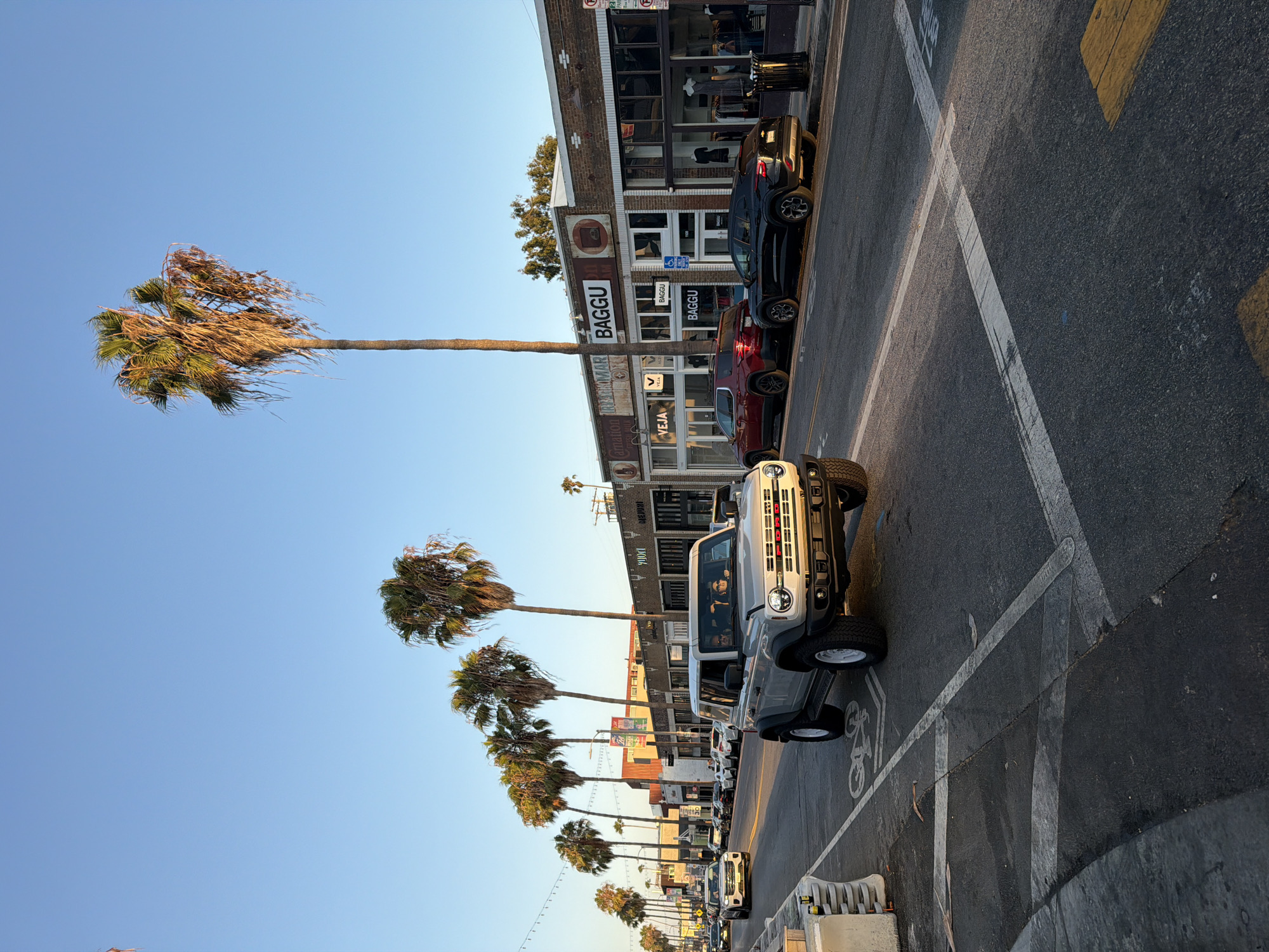 A light silver Ford Bronco drives down a street lined with palm trees and brick buildings. The buildings appear to be shops, with signs reading "VEJA" and "BAGGU." The sky is clear and blue, and the sun is setting, casting a warm glow on the scene. Several other cars are visible on the street, and some people are walking on the sidewalk. The Bronco is in the foreground, with its front end turned slightly towards the camera. The windows of the Bronco are tinted, but two people can be seen inside. A bicycle lane is painted on the road in front of the Bronco. The overall impression is one of a pleasant, urban street scene on a sunny day.