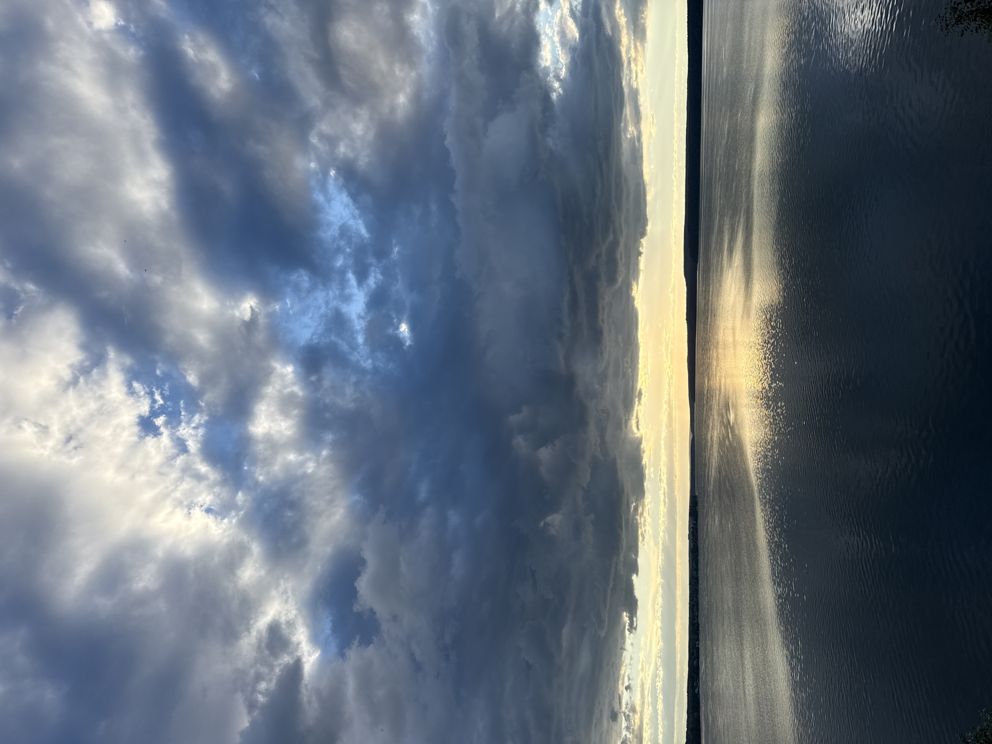 The image depicts a wide, calm body of water under a dramatic sky filled with dark, heavy clouds interspersed with brighter patches of sunlight. The horizon is marked by a dark, silhouetted treeline, indicating land. The water reflects the sky and the distant land, creating a muted, somber atmosphere. The location is identified as Votkinsk, Russia. There are no people or specific objects of note visible in the image. The scene appears to capture a moment of natural beauty, possibly before or after a storm.