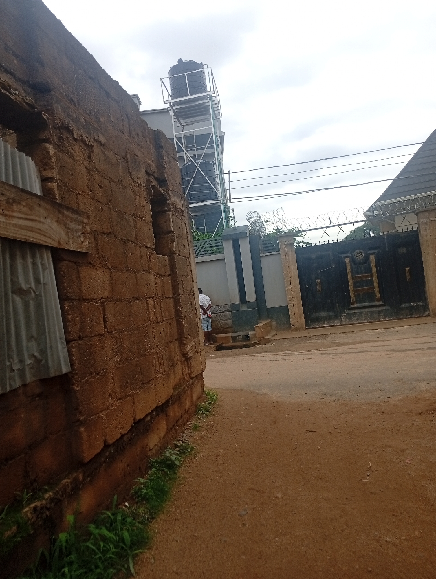 In Rafin Par, Nigeria, an overcast sky hangs above a scene dominated by a rough, earth-toned brick wall on the left. A corrugated metal sheet is affixed to the wall with wooden planks. To the right, a dirt road leads past a tall, black metal gate with decorative elements, flanked by concrete pillars. Above the gate, razor wire is stretched along a grey wall. In the background, a multi-story structure is visible with a black water tank on its roof, supported by a metal scaffolding. A person in a white shirt and shorts stands facing away from the camera near the gate.