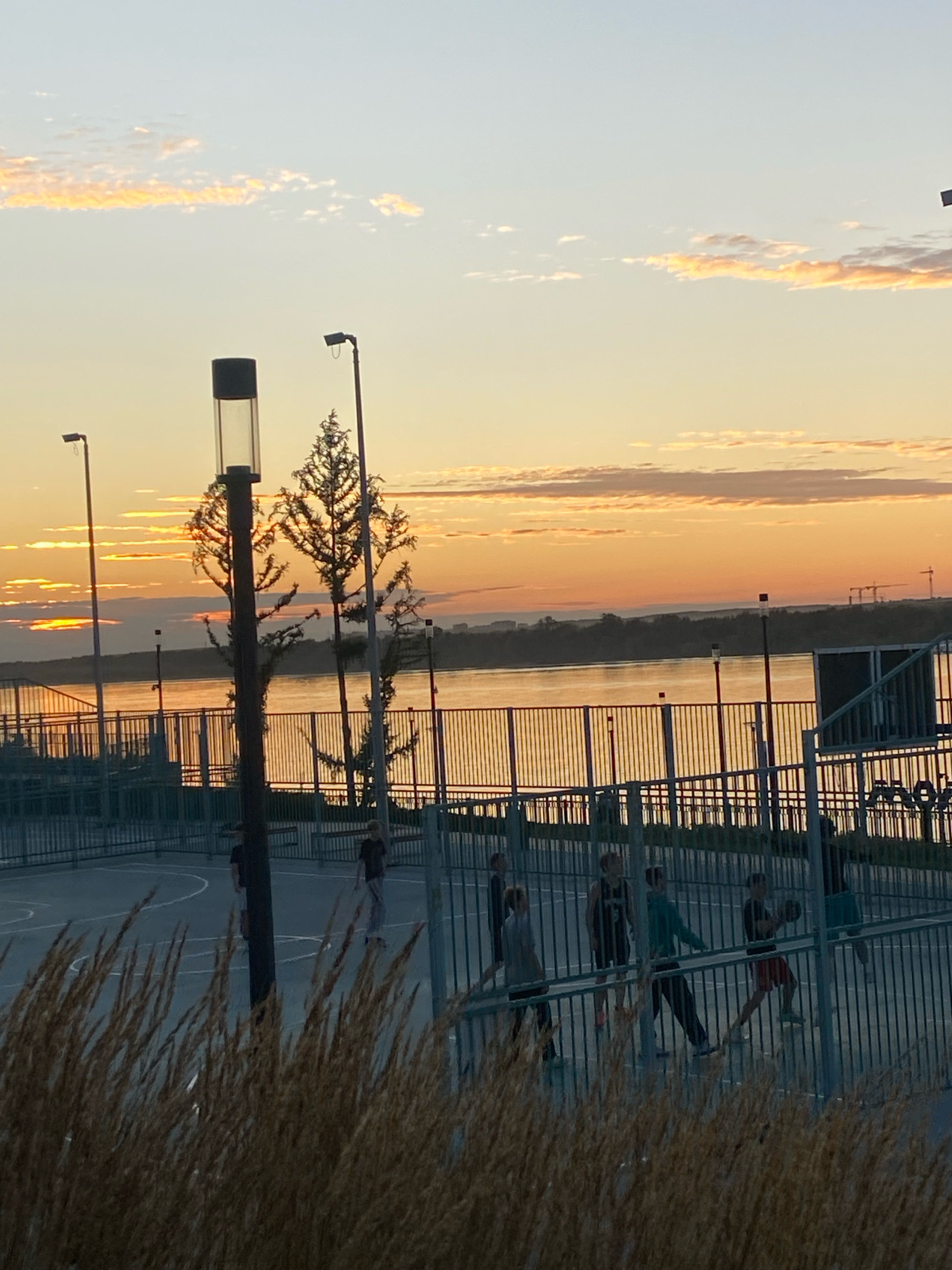 The image depicts a sunset over the Kama River in Perm, Russia. The sky is filled with soft orange and yellow hues, with scattered clouds. In the foreground, a basketball court is visible, enclosed by a metal fence. Several young people, likely teenagers, are on the court, some playing basketball and others standing around. A tall lamp post stands on the court. Beyond the court and the river, the Perm skyline is visible in the distance, with cranes suggesting ongoing construction. The overall scene captures a moment of leisure and activity at dusk by the river.