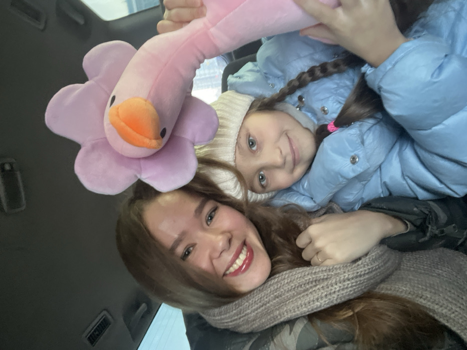 An adult female and a young female child are depicted inside a vehicle. The adult, with long brown hair and red lipstick, wears a camouflage-patterned jacket and a light brown knitted scarf, displaying a broad smile. A plain gold band is visible on her left ring finger. Seated beside her, the child, with brown hair in two braids secured with pink elastic bands, wears a light blue puffer jacket and a light cream-colored knitted beanie, exhibiting a subtle smile. The child holds a large pink plush toy, characterized by a light purple flower-shaped head and an orange beak-like protrusion. The background consists of the vehicle's gray headliner and a blurred exterior view through a window, indicating an urban setting. The precise location within Krasnoyarsk, Russia, cannot be determined from the visual information provided.