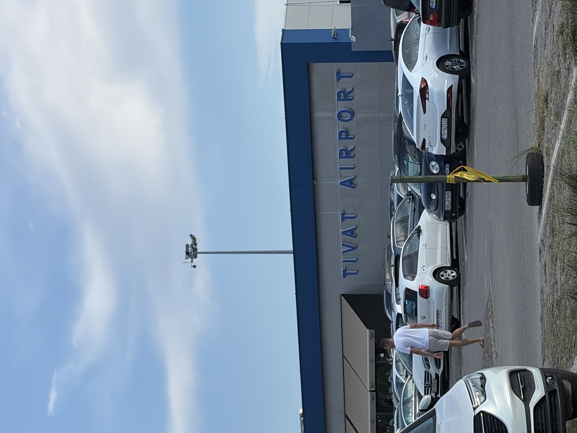 The image shows the Tivat Airport terminal in Mrčevac, Montenegro, under a partly cloudy sky. A man wearing a white t-shirt and grey shorts walks across a parking lot filled with various vehicles, including white sedans and a dark Volkswagen Beetle. The airport building features a blue upper section with "TIVAT AIRPORT" written in large blue letters. The foreground is a dirt and gravel lot with a yellow ribbon tied to a pole with a tire at its base.