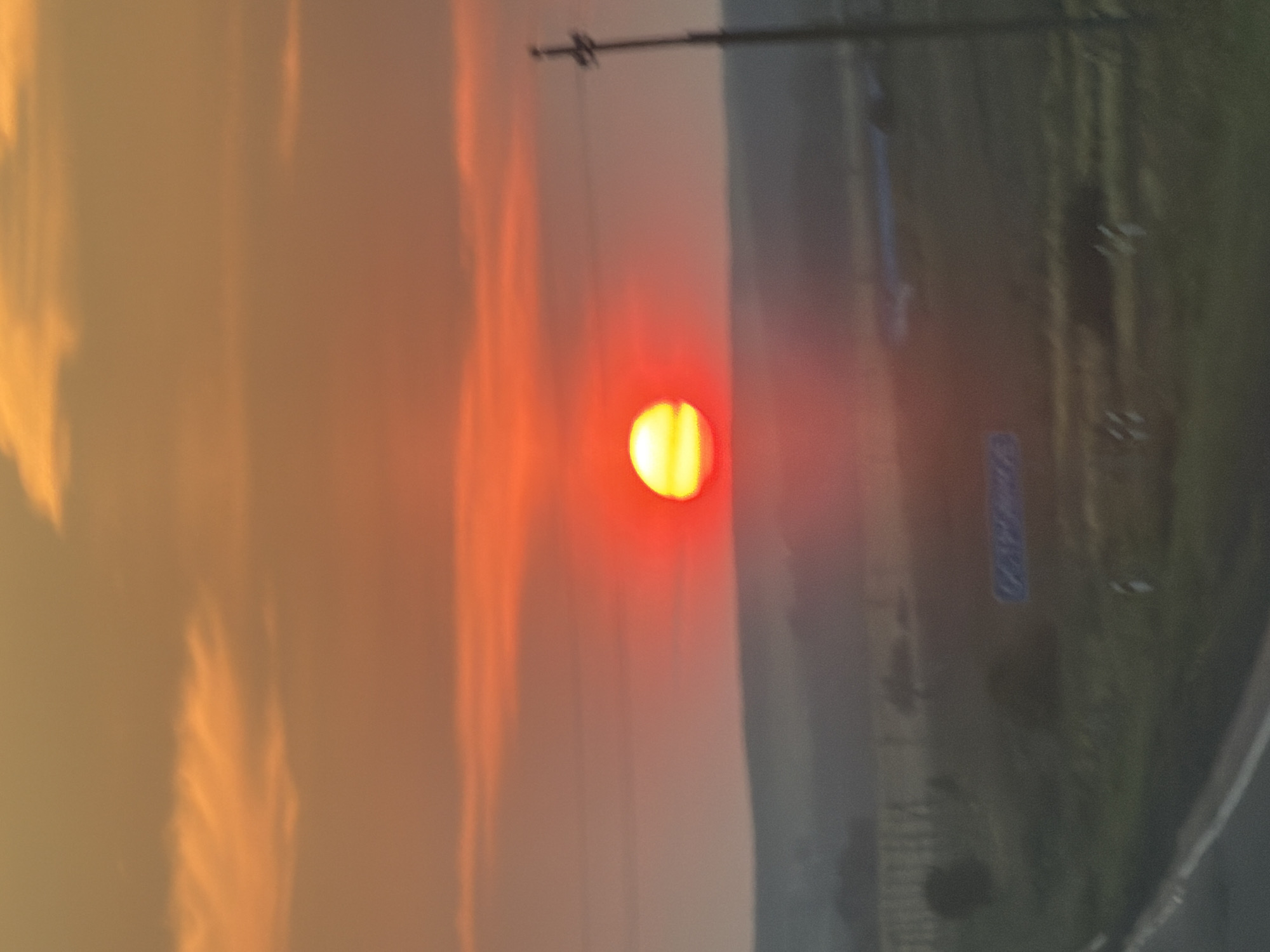 The image shows a blurry, low-angle view of a sunset over a rural landscape, likely in the city of Ostrovnoe, Russia. The sun is a bright, glowing orange orb, low on the horizon, surrounded by a hazy red and orange sky. Thin, wispy clouds are visible in the upper portion of the image. The middle ground consists of a dimly lit, undulating terrain with hints of vegetation and possibly some man-made structures. A dark, indistinct silhouette of trees or buildings is visible on the left. In the foreground, a blurred asphalt road with a white line is present. A single, dark utility pole stands on the right side of the frame, its crossbar indistinct. A faint blue sign with unreadable text is visible on the right side of the road. The overall impression is that of a scenic sunset in a relatively undeveloped area.
