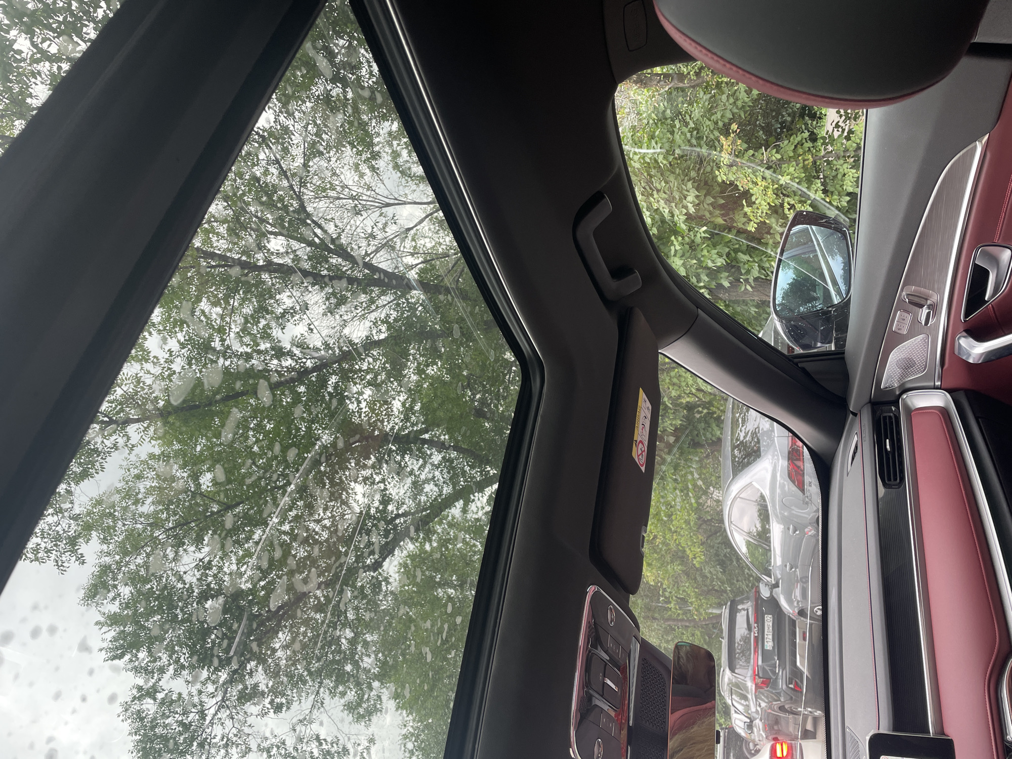 The image depicts the interior of a car, looking upwards through an open sunroof at a sky with scattered water droplets. The trees outside are lush and green, with branches visible against a cloudy sky. A black car, possibly a Kia Sportage based on the visible badge, is in traffic in front of the photographer's car. A light gray sedan is also visible in the traffic jam. The interior of the car features red and black leather seats, a metallic dashboard, and a rearview mirror. A sun visor with an airbag warning sticker is visible above the passenger side. The scene appears to be a typical urban traffic situation in Almaty, Kazakhstan, with vehicles moving through streets lined with greenery.