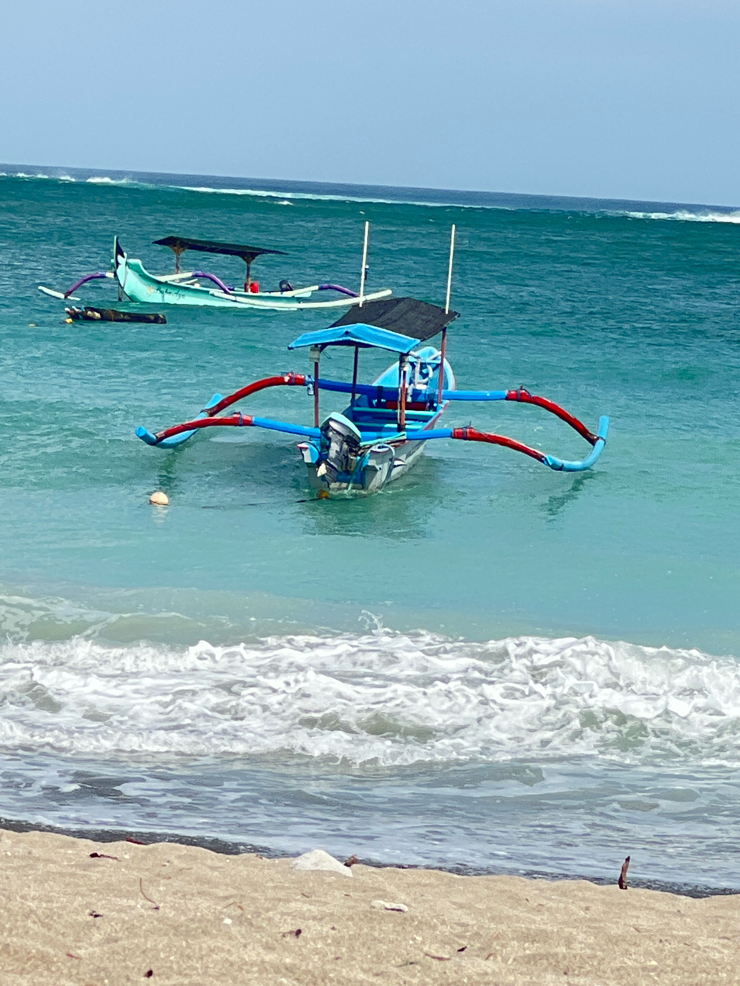 The image depicts a serene beach scene in Kuta, Indonesia, with several traditional Balinese fishing boats anchored in the turquoise waters of the ocean. In the foreground, gentle waves lap against the sandy shore, with a few pieces of driftwood scattered on the beach.
Dominating the center of the frame is a brightly colored blue fishing boat equipped with a canopy and an outboard motor. Its distinctive outriggers, painted in red and blue, extend outwards, providing stability. A small, orange buoy floats nearby, tethered to an unseen anchor.
In the background, another light green Balinese fishing boat is visible, also with an outrigger and canopy. Further in the distance, the ocean stretches out to the horizon, with faint whitecaps suggesting the presence of larger waves. The sky above is a clear, pale blue, indicating fair weather. The overall atmosphere is one of tranquility and natural beauty, characteristic of the Kuta coastline.