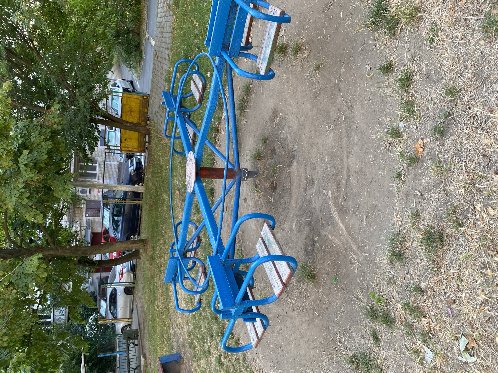 In Chornomorsk, Ukraine, a well-used blue metal merry-go-round with wooden seats stands in a dirt playground area. The merry-go-round is equipped with six seats, each with a protective blue U-shaped bar. Behind the playground, several parked cars, including a silver sedan and a dark blue sedan, are visible next to a multi-story building with balconies. Trees with green foliage are scattered throughout the scene, providing shade. The ground is a mix of dirt, dry grass, and scattered leaves. The overall impression is of a typical urban playground in a residential area.