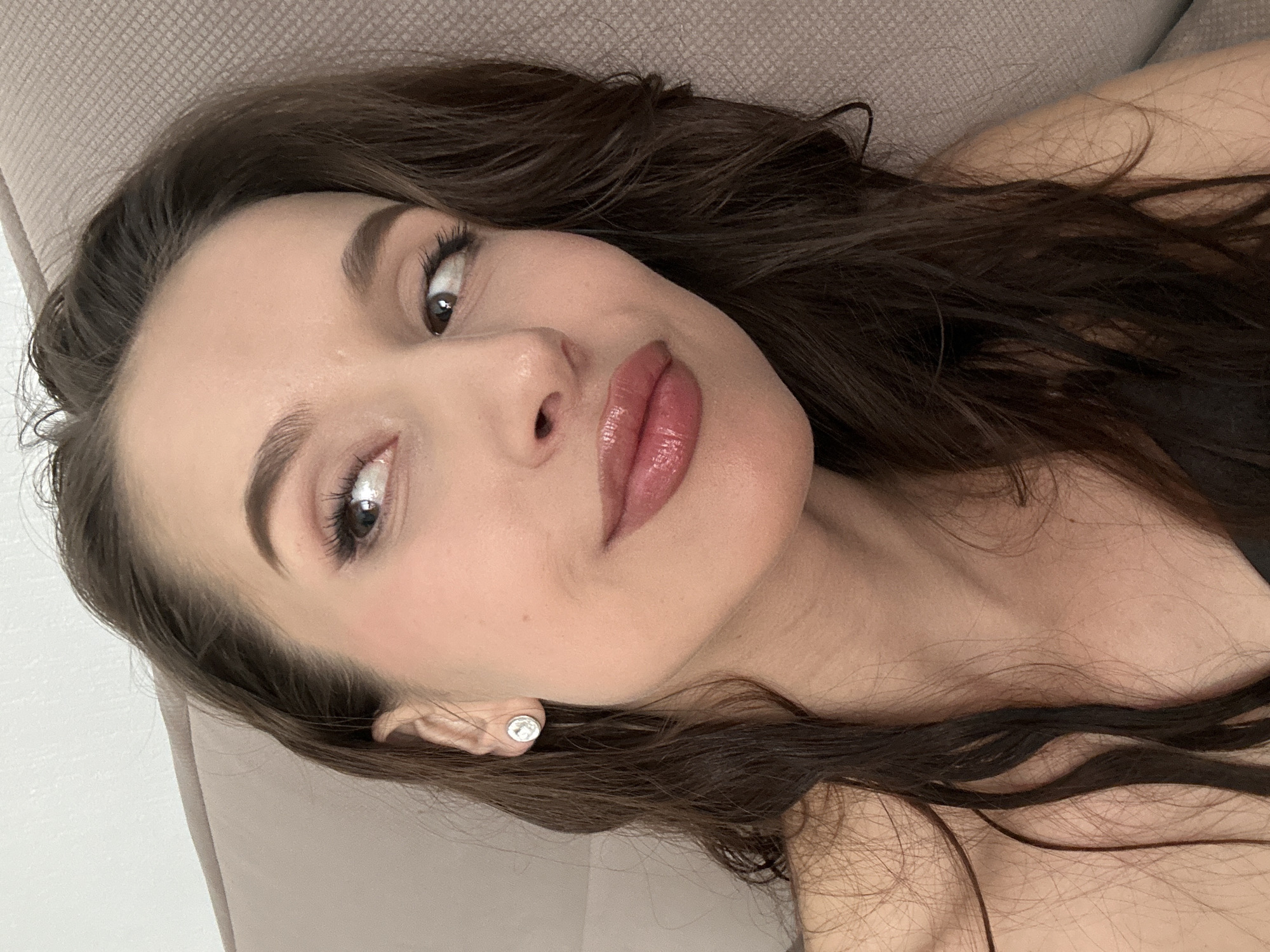 A close-up selfie captures a young woman with dark brown hair. Her hair is styled in loose waves. She has fair skin, defined eyebrows, and glossy pink lips. She is wearing a small, round earring in her left ear. The background consists of a neutral-colored, possibly beige, textured surface. The image provides no information about the location in Yekaterinburg, Russia, or any actions, interactions, or subjects in the environment.