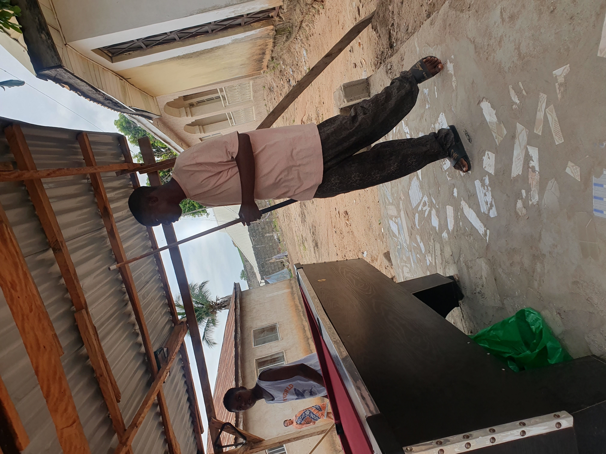 An outdoor scene in Lokoja, Nigeria, shows two young males and one female near a pool table. The younger male in the foreground stands to the left of a pool table, looking at the camera. He wears a white tank top with a graphic design and dark pants. The other male, in the mid-ground, is seen from the side and back, holding a pool cue and wearing a light pink t-shirt and patterned dark pants. A woman in the background wears a patterned wrap skirt and top. The environment includes a covered outdoor area with a corrugated metal roof supported by wooden beams, a pool table, and a sandy, uneven ground with scattered building materials. In the distance, there are simple residential buildings and some vegetation. A green plastic bag is visible under the pool table.