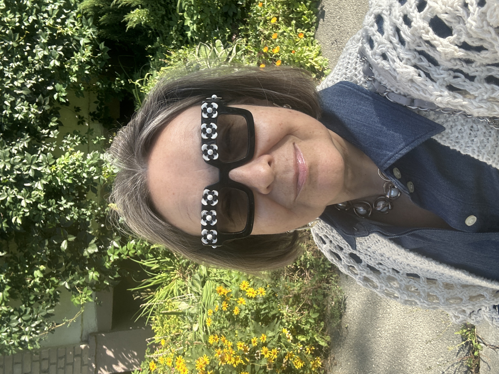 A middle-aged woman, with grey hair and fair skin, is pictured in a medium shot, looking at the camera. She is wearing large, square-shaped black sunglasses decorated with white and black floral patterns on the top of the frames. She has on a denim shirt and a cream-colored, open-knit cardigan. A chunky silver necklace with pearl-like accents is visible. The background consists of lush green foliage and yellow flowers, suggesting an outdoor setting in a garden or park. The lighting indicates it is daytime, with sunlight casting shadows on the ground.