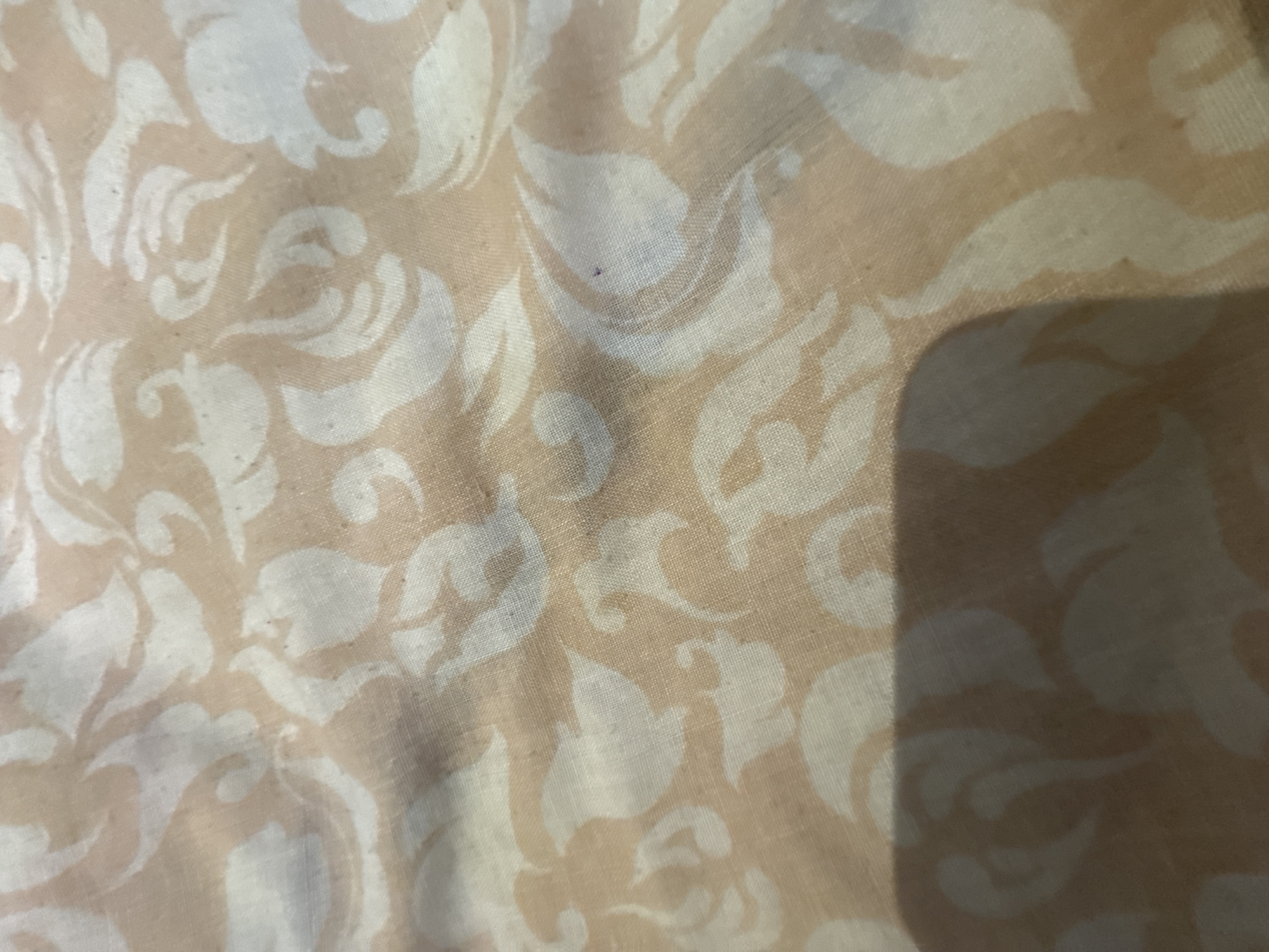 The image shows a close-up view of a fabric with a floral pattern in shades of peach and off-white. The fabric appears to be a thin material, possibly a curtain or a piece of clothing, with a slightly textured surface. The pattern consists of stylized leaves and swirling shapes. The lighting suggests an indoor environment, with a shadow cast across the lower portion of the image, obscuring some of the pattern. There is no information in the image that can be used to determine its location in the city of Poltava, Ukraine.