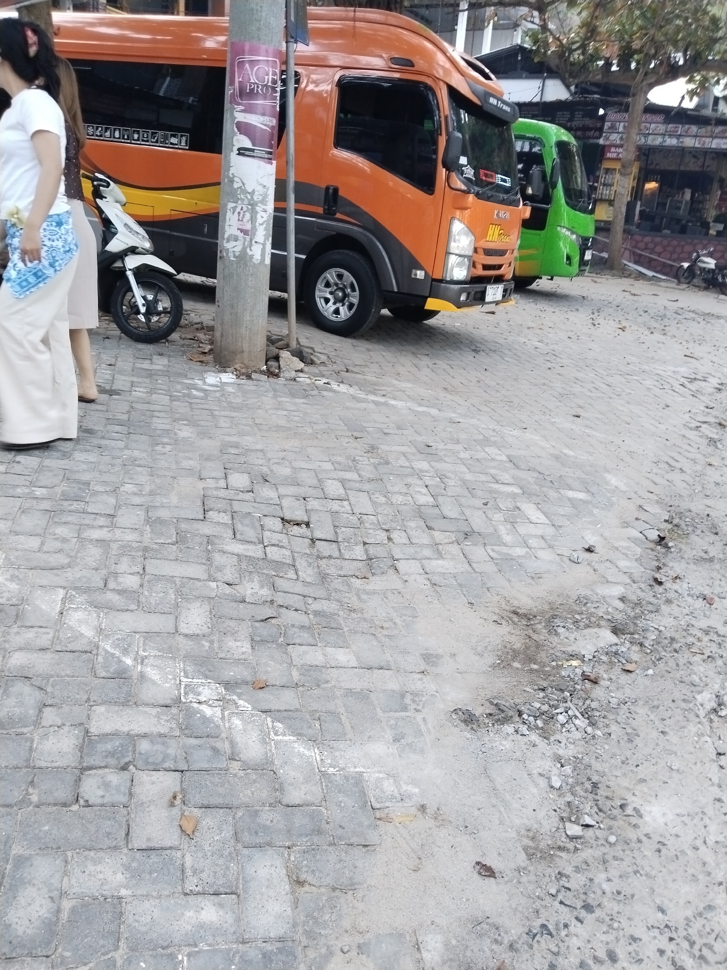 In Prigi, Indonesia, an orange and yellow tour bus is parked on a cobblestone street. A green bus is parked behind it. A white scooter is parked in front of the orange bus. A woman wearing a white shirt and beige pants stands on the left side of the frame. The street is lined with shops and trees. The sky is overcast.