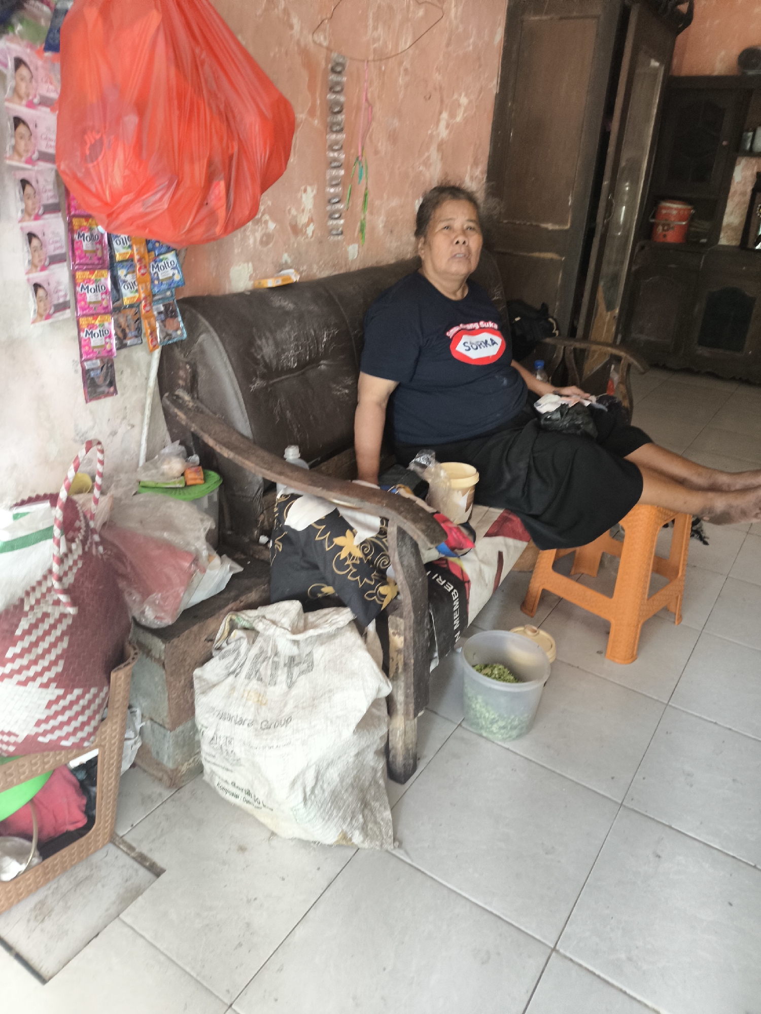 An older woman sits on a sofa in a sparsely furnished room. She is wearing a dark blue t-shirt with a circular logo on it. She is wearing a black skirt and is barefoot. There is a large red plastic bag hanging on the wall behind her, and a shelf with various items is to her right. In front of her, on the floor, are several objects including a large white bag, a woven red and white bag, and a clear plastic container filled with chopped green vegetables. The room appears to be a residential interior. The location of the scene is identified as Citrodiwangsan, Indonesia.