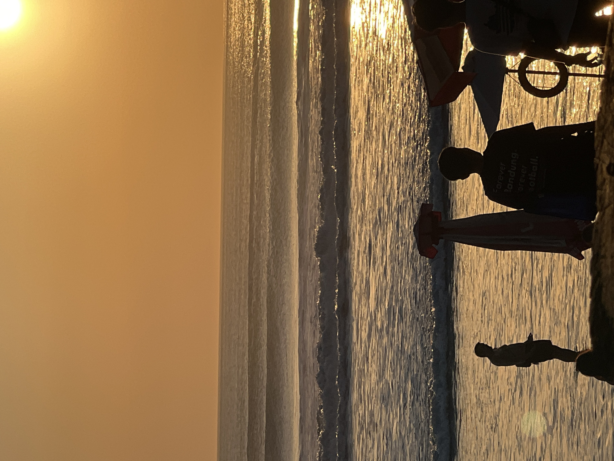 The image depicts a sunset scene at Kuta Beach, Indonesia. Silhouetted figures stand on the shore as waves roll in, reflecting the golden light of the setting sun. In the foreground, several people are visible, some standing in the shallow water, while others are near beach umbrellas and what appears to be a life preserver. The sky is a warm orange hue, and the ocean stretches out to the horizon. The overall atmosphere is serene and tranquil.