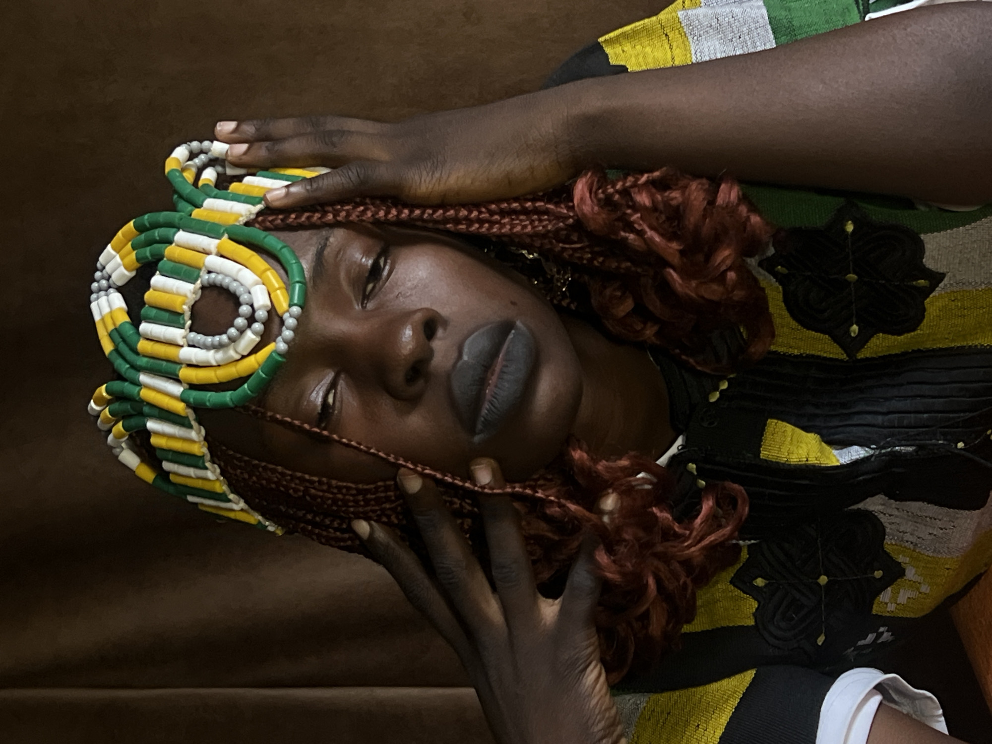 The image is a close-up portrait of a Black woman wearing a beaded headdress and a patterned top. The headdress consists of a circular arrangement of cylindrical beads in green, yellow, white, and gray. Her hair is styled in braids that frame her face and extend down. She is wearing dark lipstick and her eyes are looking downwards. Her hands are raised, with fingers gently touching her head and face. The background is a dark, textured brown, suggesting a studio setting or a dimly lit room. The woman is likely within the city of Jos, Nigeria, due to the cultural context suggested by the traditional headdress and attire, though the image itself does not provide explicit geographical markers.