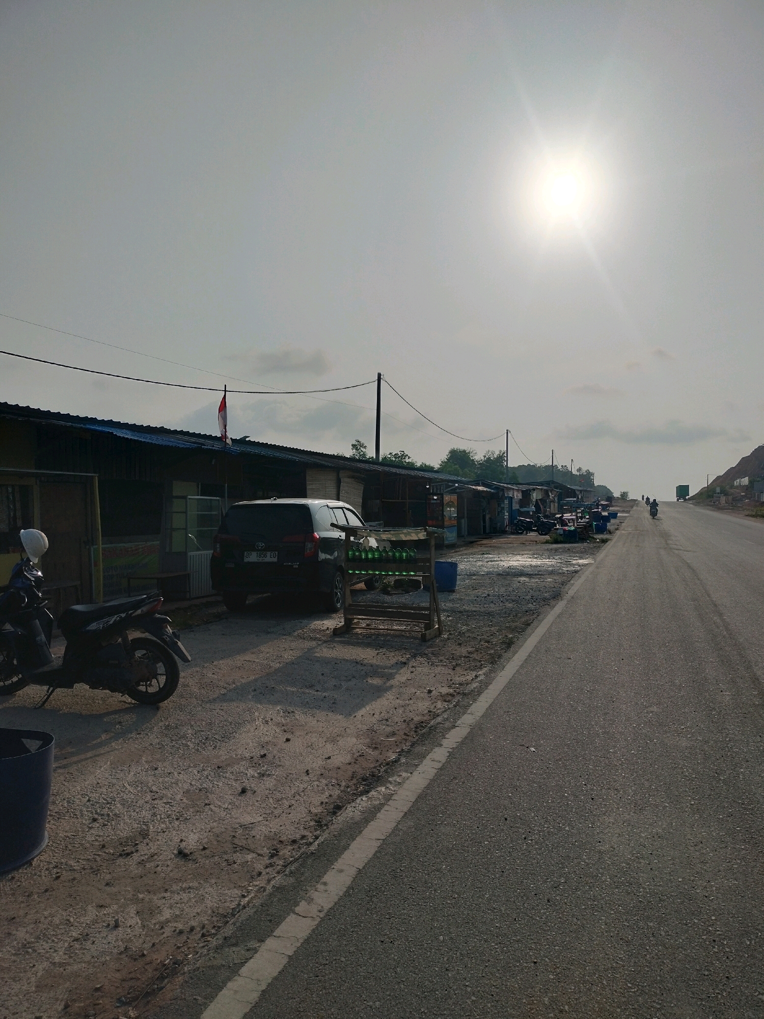 The image captures a roadside scene in Batam, Indonesia, likely a commercial area along a highway. In the foreground, a dark-colored motorcycle is parked on a gravelly shoulder. To its left, a dark blue cylindrical bin is visible. A black SUV is parked further down the road, in front of a row of low-rise buildings with corrugated metal roofs. These buildings appear to be small shops or stalls, with some signage and display racks visible. A red and white flag flies from one of the buildings. The road itself is paved, with a white line marking the edge. The sun is high in a hazy, overcast sky, casting a bright glare. In the distance, the road curves to the right, with a hillside on the far right side. Several motorcycles and a truck are visible on the road in the distance, indicating ongoing traffic. The environment appears to be somewhat arid, with sparse vegetation visible on the hillside. The overall impression is that of a typical roadside service area or collection of small businesses operating along a major thoroughfare in Batam.