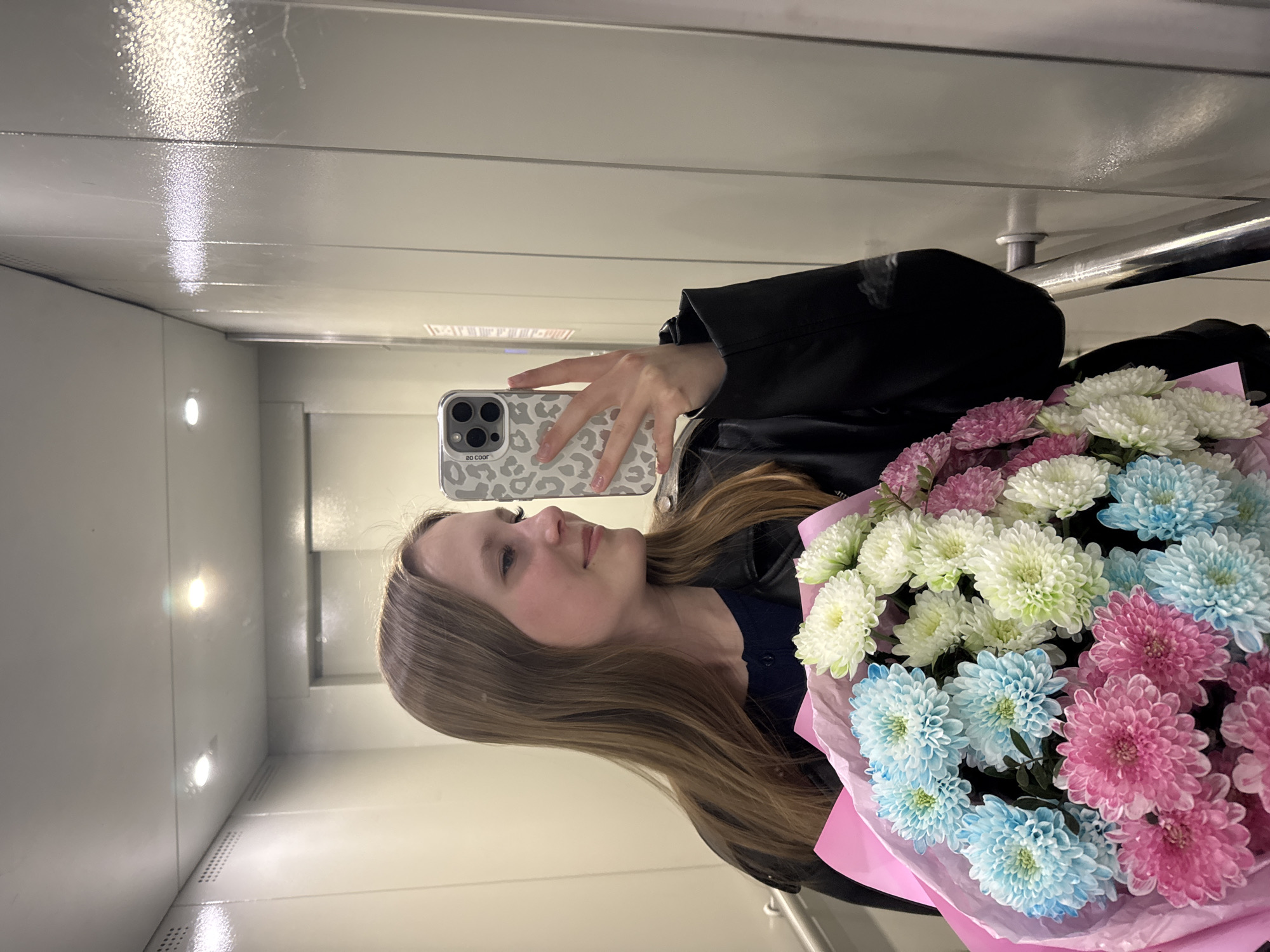 A person is taking a selfie in what appears to be an elevator. The individual is holding a large bouquet of flowers in the foreground, consisting of white, light blue, and pink chrysanthemums wrapped in pink paper. The person is wearing a black jacket. Their phone, with a grey animal print case, is visible in their hand as they take the photo. The elevator interior is white, with a metallic handrail visible in the background. Based on prior information, the location is Chaikovsky, Russia.