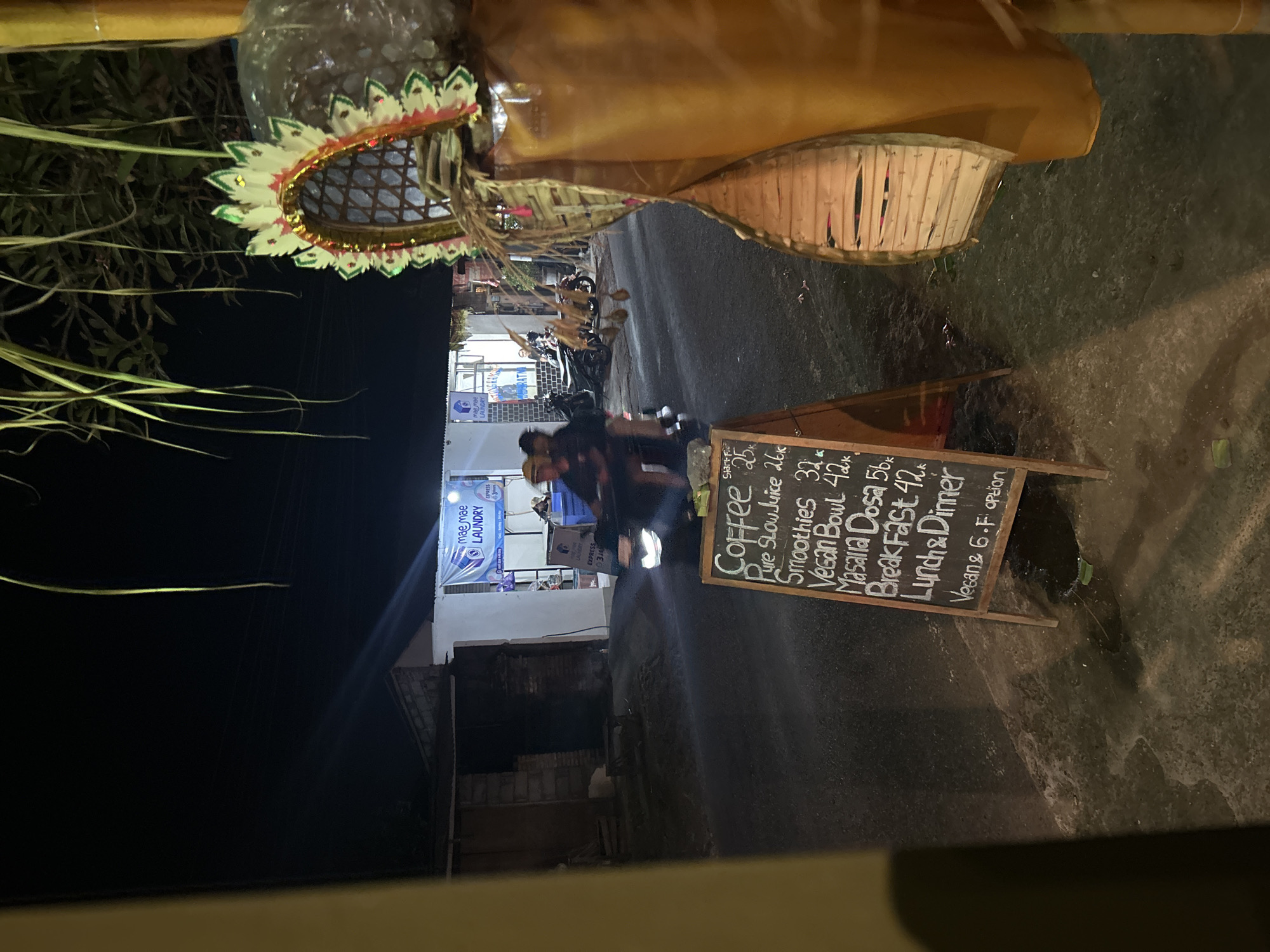 This media file captures a nighttime scene on a street in Ubud, Indonesia. The focus is on a chalkboard menu positioned in the foreground, displaying food and drink items. Items listed include "Coffee" for 25, "Pure Slow Juice" for 26k, "Smoothies" for 32, "Vegan Bowl" for 42k, "Masalla Dosa" for 56k, and "Breakfast" for 42. The menu also mentions "Lunch & Dinner" and notes "Vegan & G.F option".
Behind the menu, a building with a "mae mae LAUNDRY EXPRESS" sign is visible. Several motorcycles are parked in front of the building, suggesting a local establishment. The lighting indicates it is nighttime, with artificial lights illuminating the scene. The ground appears to be wet, possibly from recent rain, reflecting the lights. The overall atmosphere is casual and appears to be a street-side eatery or shop. The presence of the decorative elements at the top of the image, including woven bamboo and colorful adornments, suggests a cultural or festive context, common in Indonesia. The image is oriented vertically, with the top of the frame showing a partial view of decorative elements and foliage.