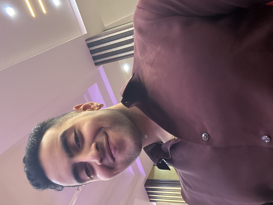 A smiling man with dark hair and a neatly trimmed beard poses indoors, his head slightly tilted. He's wearing a burgundy dress shirt with visible buttons. The background features a white ceiling with recessed lighting and a strip of silver and black patterned material. The lighting is soft, creating a warm and inviting atmosphere.