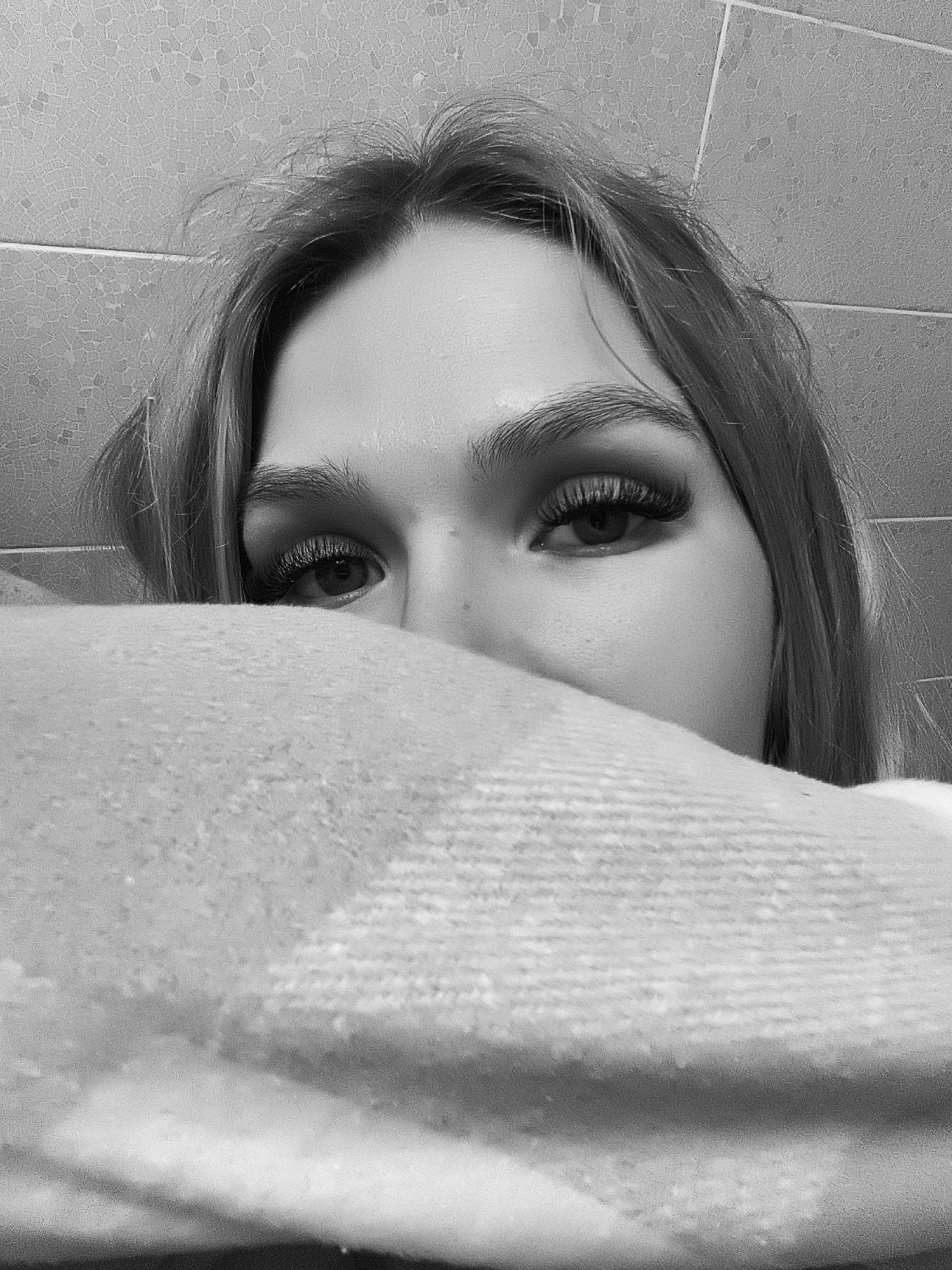 A close-up black and white photograph shows a person's face partially obscured by a soft, textured blanket or fabric. Only the upper part of the face is visible, specifically the forehead, eyes, eyebrows, and the bridge of the nose. The person has long eyelashes and defined eyebrows. The background appears to be a tiled wall. The image is out of context and therefore no further information about Izhevsk, Russia, actions, interactions or specifics regarding the environment can be accurately described.
