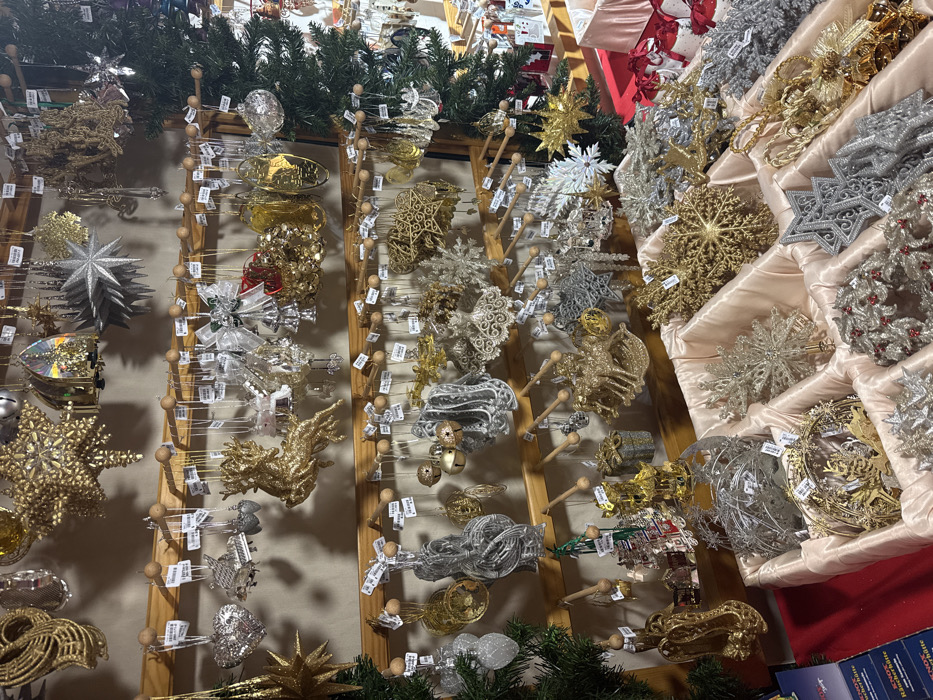 Rows of Christmas ornaments in gold, silver, and clear designs are displayed on wooden stands and in cushioned trays. The ornaments vary in shape and material, including stars, snowflakes, and delicate wire structures. Many are embellished with glitter or intricate cutouts. Greenery is placed on top and bottom of displays. Price tags dangle from the ornaments.
