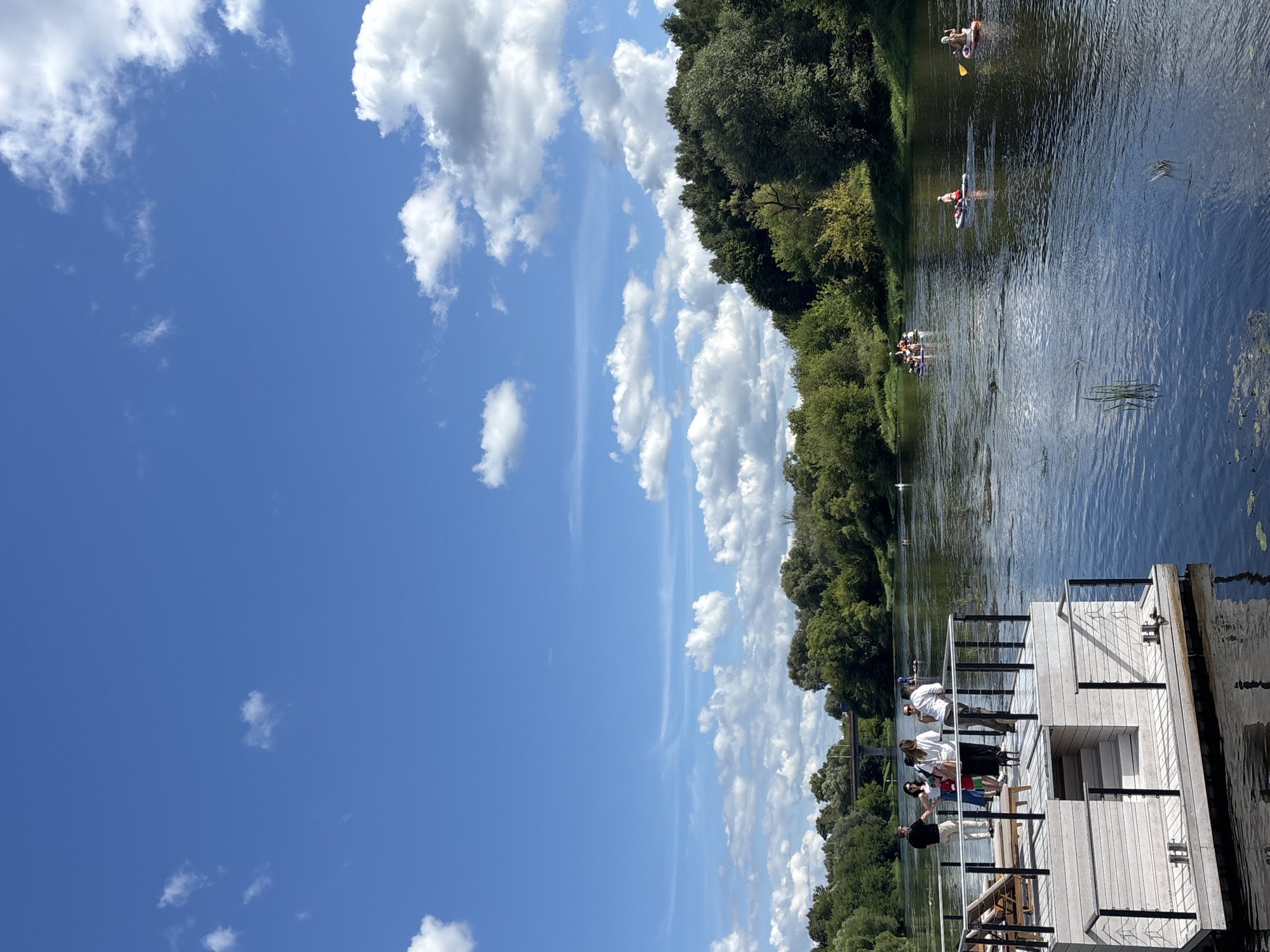 This image captures a sunny day by the Moskva River in Zvenigorod, Russia. In the foreground, a modern, multi-level wooden deck juts out over the water, with several people standing and conversing on its upper tier. The deck is furnished with lounge chairs and has black metal railings. The river itself is calm, with a few water lilies visible near the edge.
Further out on the river, individuals are engaged in water activities. Several people are paddleboarding, with one person paddling an orange board and another on a white board. A group is clustered together on a larger inflatable raft or boat. In the background, the riverbanks are lined with dense green trees, and a bridge can be seen spanning the river in the distance. The sky is a vibrant blue, dotted with fluffy white clouds, indicative of pleasant weather. The scene conveys a sense of leisure and outdoor recreation on a summer day.