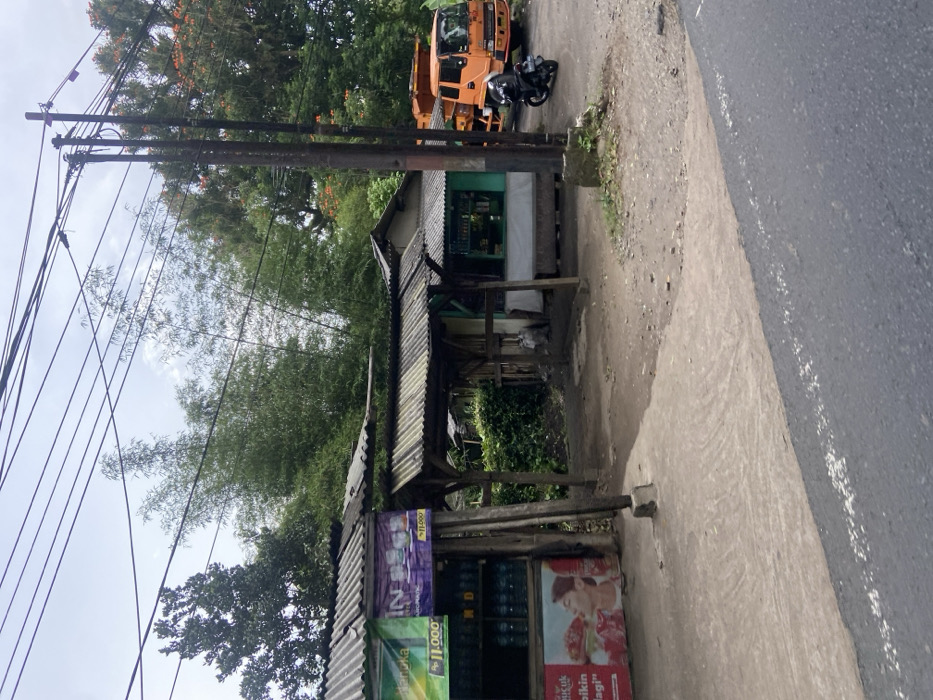The scene shows a street in Cipatat, Indonesia (latitude: -6.794, longitude: 107.442). On the left side of the road is a small store with a corrugated metal roof. The store has a sign in the front advertising "Rp. 11.000" and "Rp. 11.000." The sign also contains the word "Tanjungka" and a purple and white sign with the word "IN" on it. Behind the store is a wooden frame structure with a wooden slatted roof. On the opposite side of the street is a large, orange truck with a small motorcycle parked in front of it. The truck has a "00001" sticker on its side. The truck is partially obscured by a cluster of trees on the right side of the frame. Behind the store, green foliage is visible. There are several power lines running overhead. The scene is lit by overcast sky.