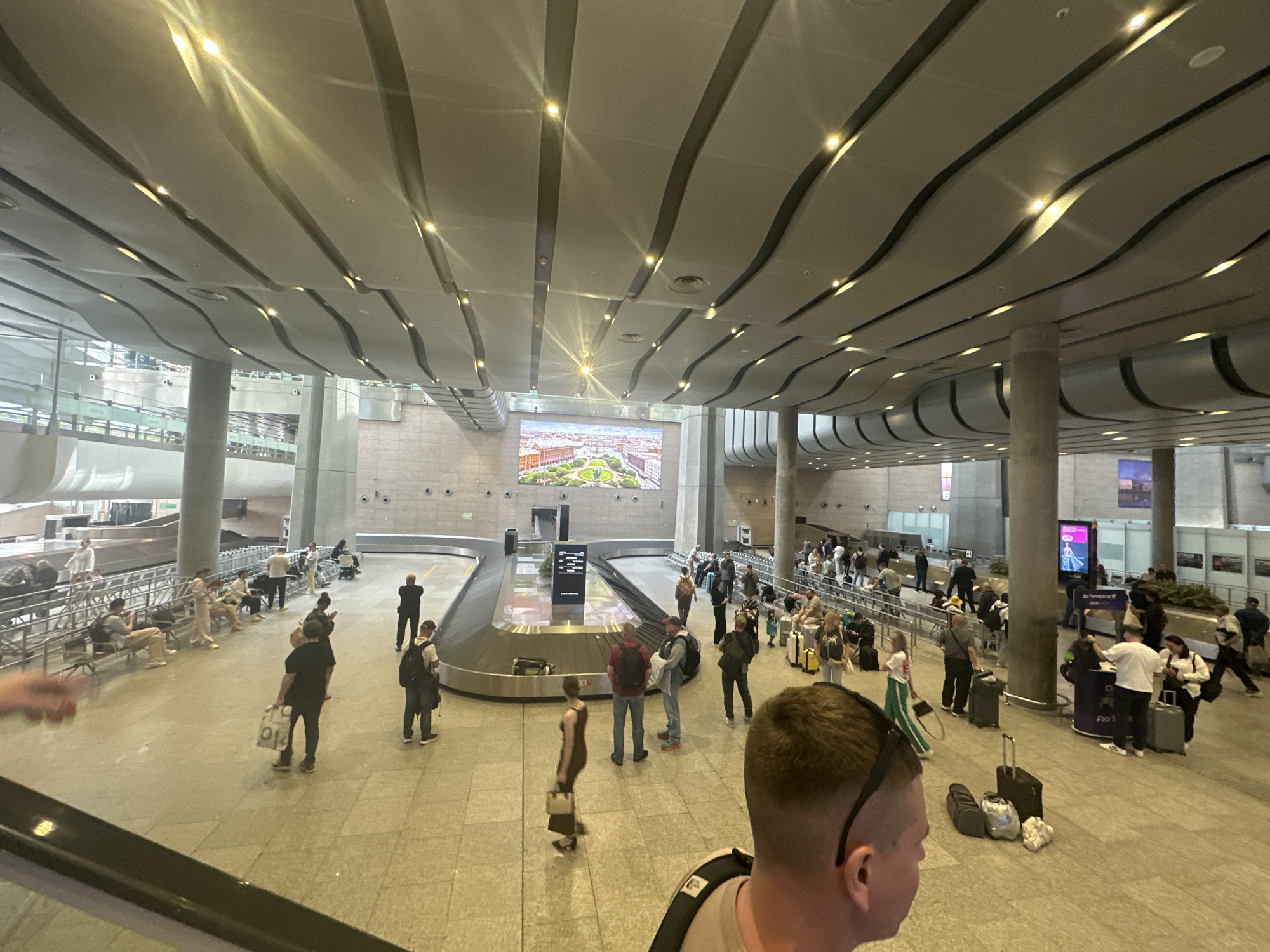 The image shows the interior of Pulkovo Airport in Saint Petersburg, Russia. The scene is likely a baggage claim area, with a central carousel surrounded by people waiting for their luggage. Many individuals are standing, while others are seated on benches. Luggage carts and suitcases are visible around the perimeter. A large screen displays an image of Saint Petersburg, likely intended to welcome visitors. The environment is modern and spacious, characterized by a high ceiling with a distinctive wavy design and recessed lighting. Pillars are spaced throughout the area.