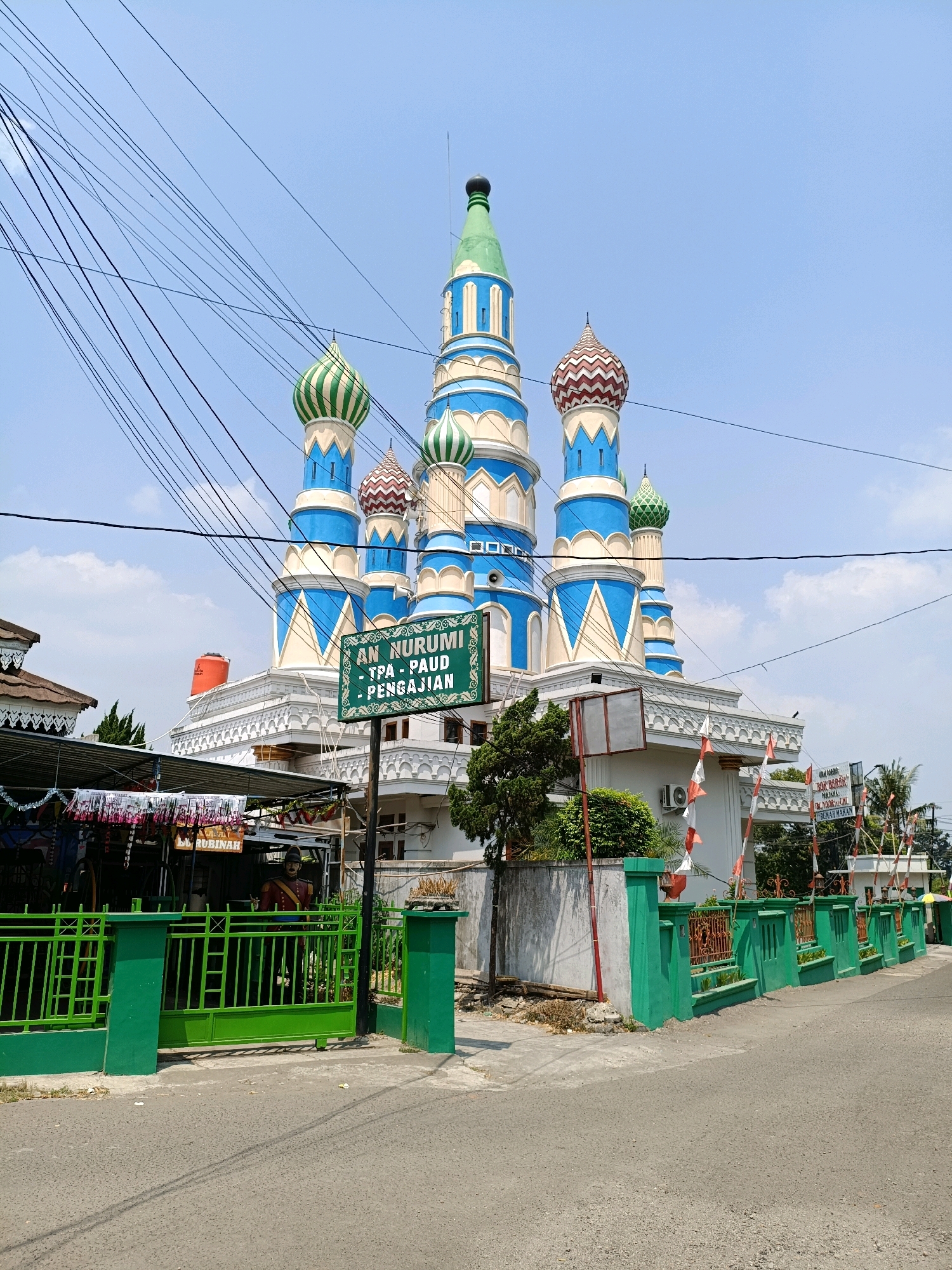A large, ornate building with multiple colorful domes, resembling a mosque with Russian architectural influences, stands under a bright blue sky with scattered clouds. The building features a prominent green and blue color scheme with white accents, and its domes are topped with green spires. In front of the building, a green fence encloses a yard. A sign in front of the building reads "AN NURUMIH - TPA - PAUD - PENGAJIAN." Several utility wires crisscross the sky above the building. To the left of the main structure, a smaller building with a white, peaked roof is visible. The scene appears to be a street in a residential or semi-commercial area, with an asphalt road in the foreground and some trees and vegetation around. The overall impression is that of a cultural or religious institution.