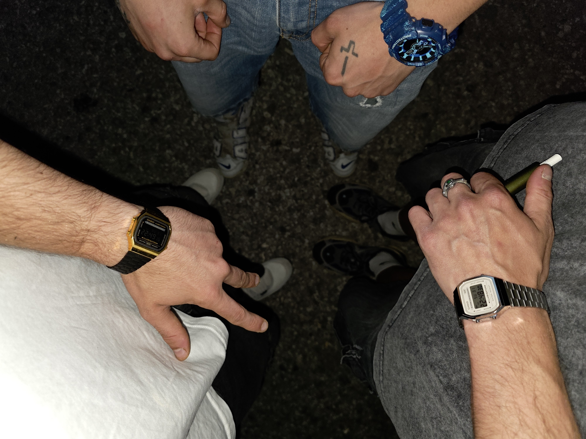 Four people are standing in a circle outdoors at night. Three are wearing wristwatches: a gold Casio on the left, a blue G-Shock at the top, and a silver Casio on the right. The person on the right is holding a vaping device. The location is unknown, no identifiable place is recognizable, there are no other objects or actions visible.