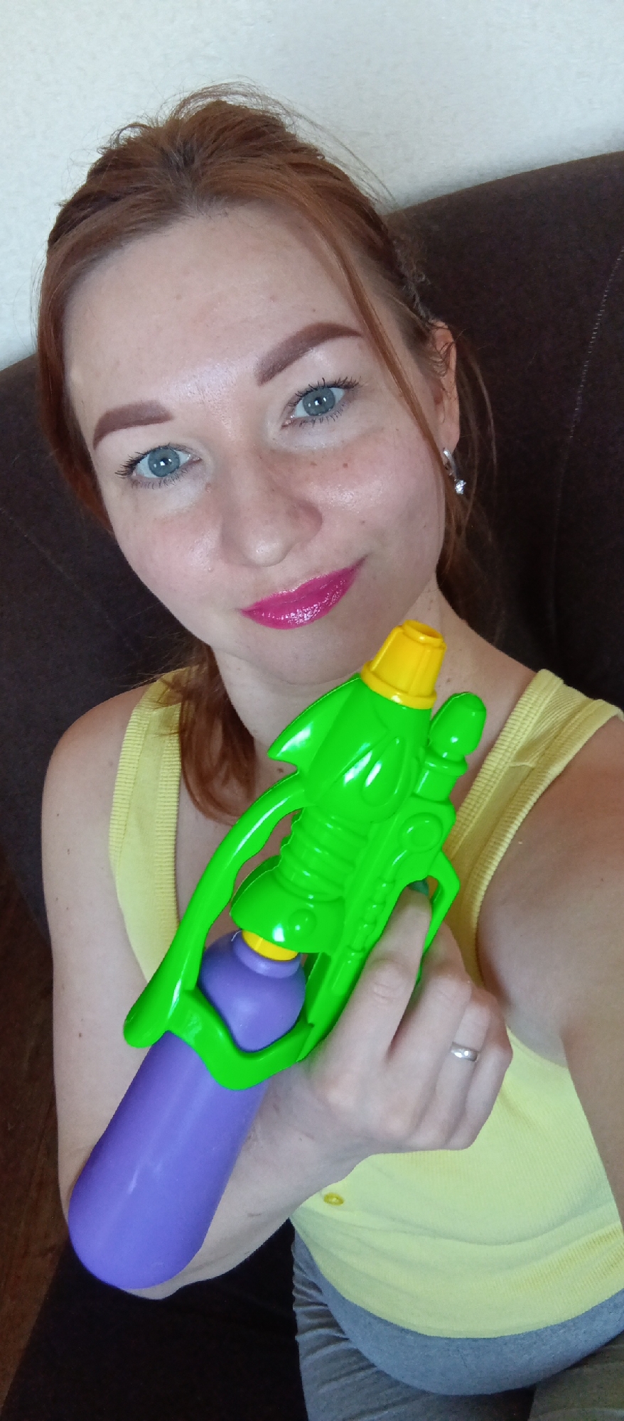 A woman with red hair and blue eyes, wearing a yellow tank top, is holding a green and purple water gun. She has bright pink lipstick on and is wearing a silver ring on her left ring finger and silver earrings. She is indoors, sitting on a brown couch, with a light-colored wall behind her. The city and location are unknown.