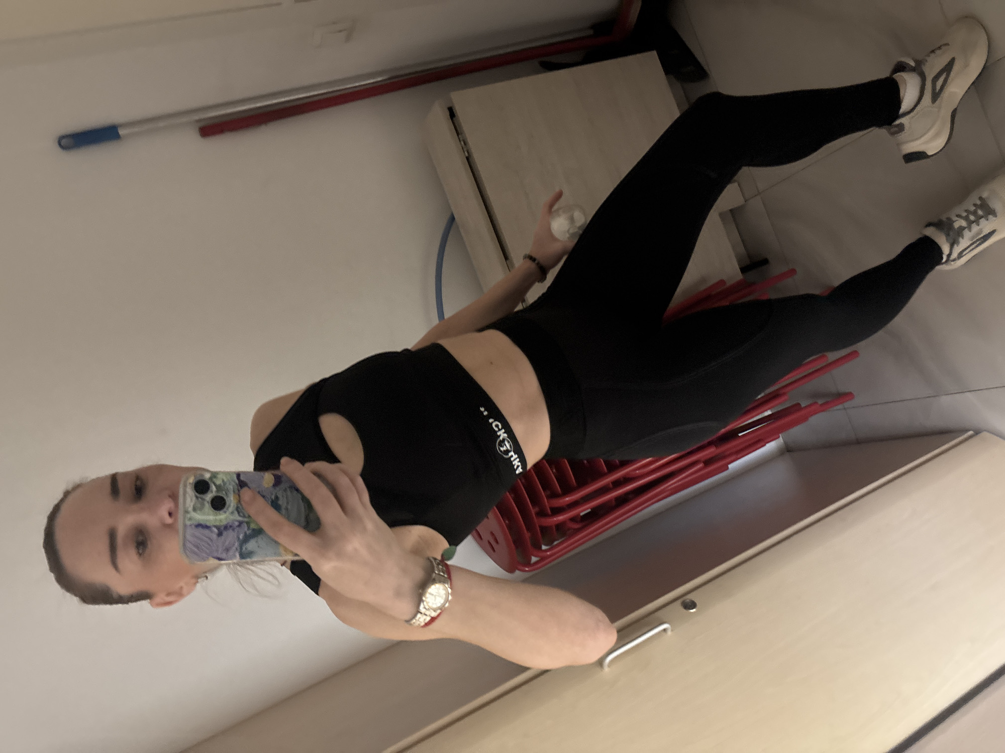 A young woman with her hair pulled back takes a mirror selfie indoors. She is wearing a black sports bra and black leggings, with white and grey athletic shoes. A silver watch is visible on her left wrist. She holds a smartphone with a colorful case in her right hand to take the photo. In the background, there are stacked red chairs, a wooden cabinet, a broom with a blue handle, and a partially visible mop. The flooring appears to be tiled. The overall environment suggests a domestic or utilitarian space. The city of Unknown, Russia is not discernible from the image.
