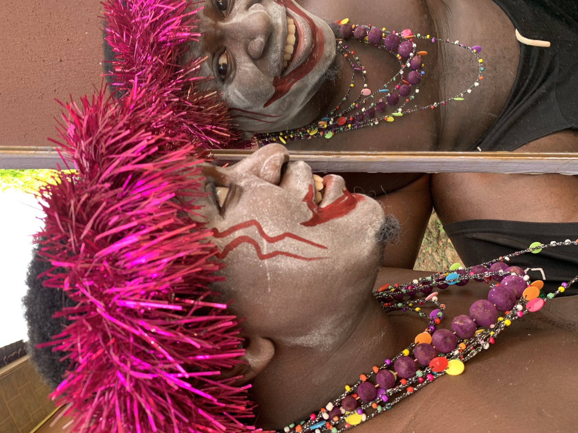 A digitally altered close-up image shows a person in costume with a mirrored reflection, appearing as two distinct faces. The faces are painted with white makeup and red markings around the mouth and on the forehead. The person is wearing a bright pink tinsel headpiece and a beaded necklace with large purple beads and colorful accents. The person is wearing a black garment that appears to be a tank top or a similar piece of clothing. The scene is cropped closely around the subjects.
It is impossible to discern the specific location in the city of Akwanga, Nigeria from the information provided.