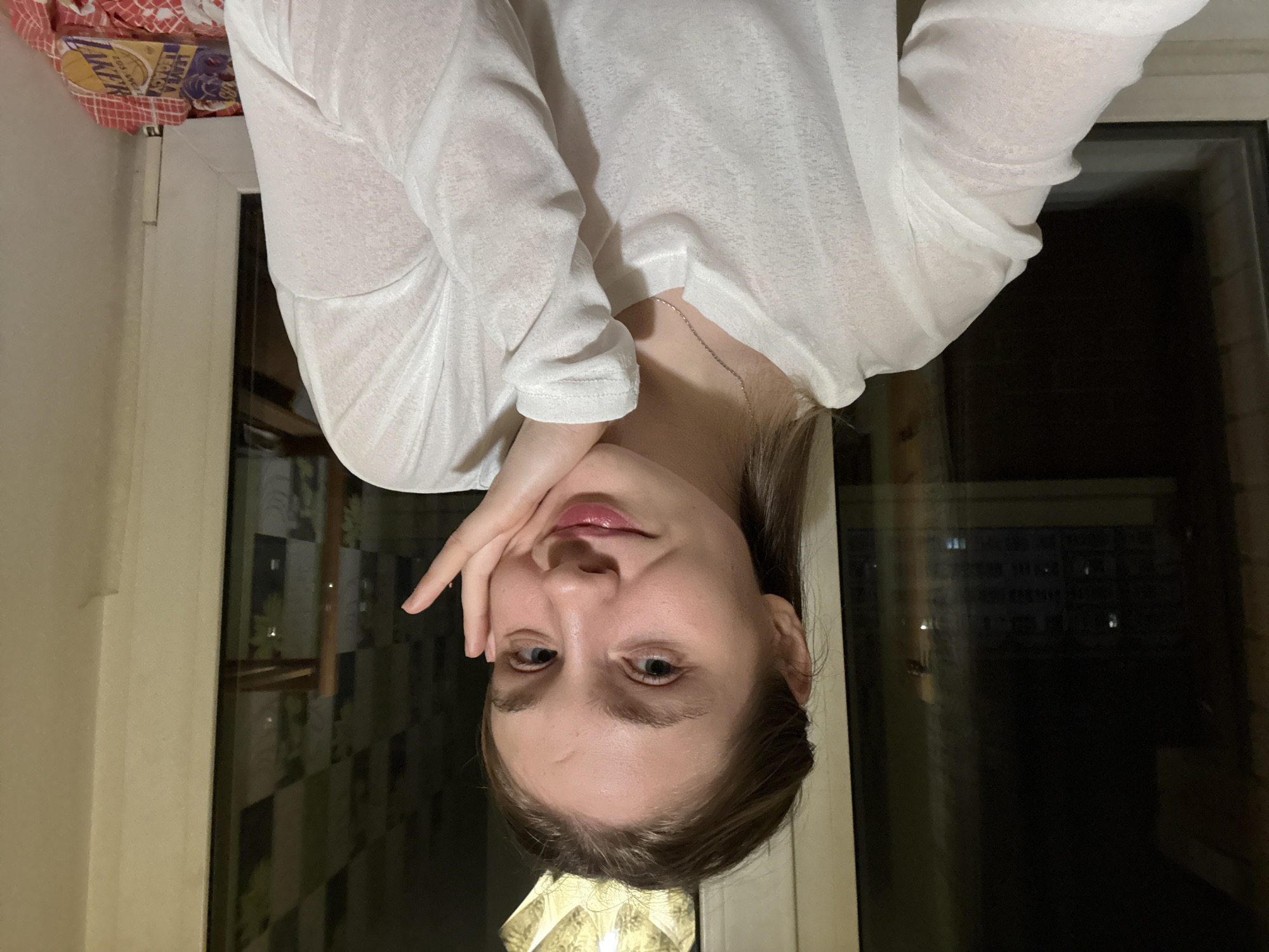 A young woman with fair skin, light brown hair, and blue eyes is positioned upside down in the frame. She wears a white, long-sleeved shirt and a delicate silver necklace. Her right hand is gently resting on her cheek. She appears to be looking directly at the camera.
Behind her is a window showing an outdoor nighttime scene. The reflection in the glass indicates the presence of other buildings, suggesting an urban environment. Based on location information, this could be Izhevsk, Russia.
On the top left is a blurred corner of what looks like a patterned bed covering, adding a domestic element to the scene.