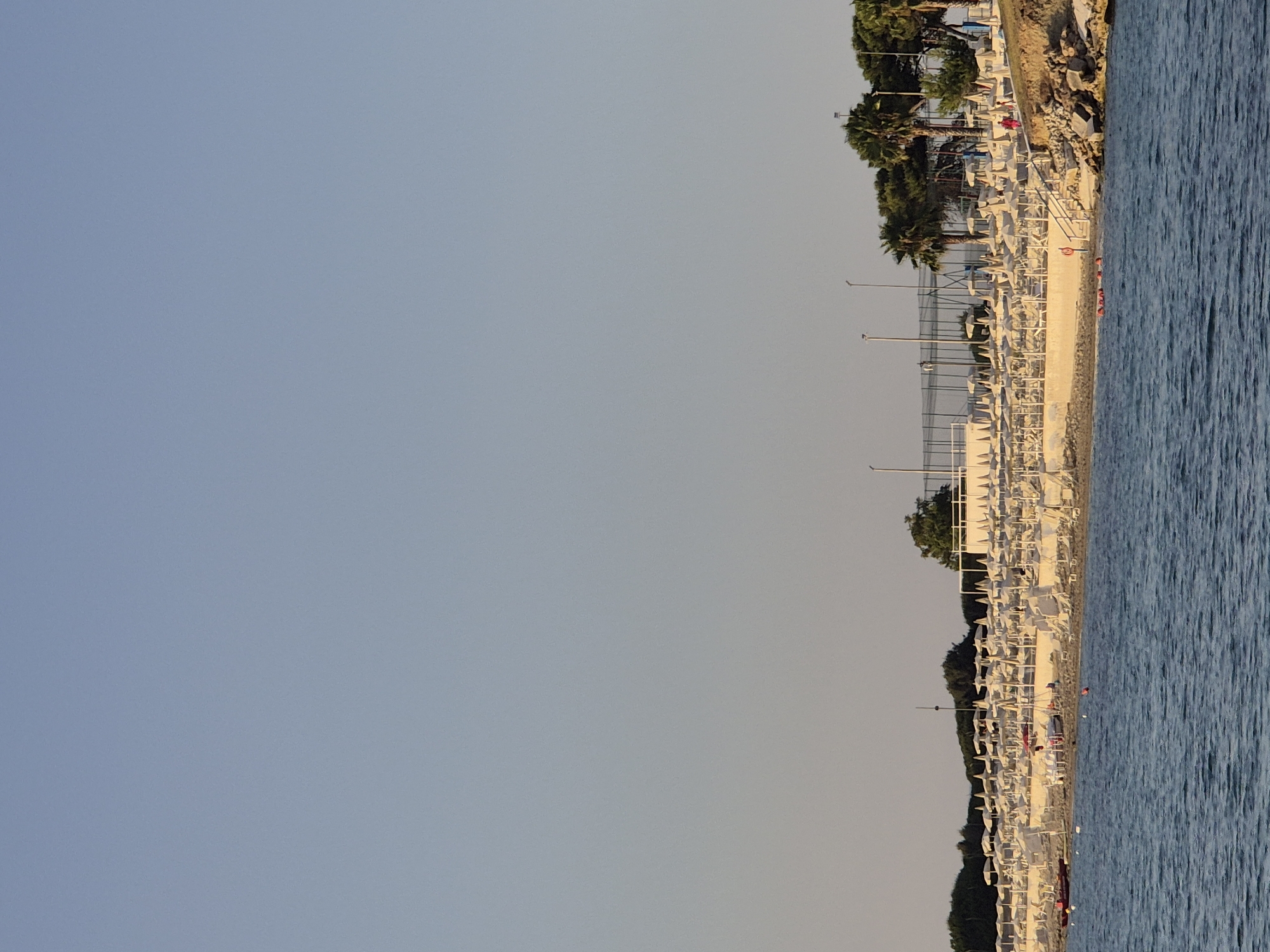 The image captures a view of a beach area in Taranto, Italy, characterized by a meticulously arranged series of beach chairs and umbrellas. The scene is set against a backdrop of a clear, light blue sky. The beach itself is dotted with numerous white lounge chairs and parasols, suggesting a well-maintained resort or public beach. In the foreground, the blue water of the sea is visible, with small ripples indicating a calm condition. A few buoys are floating in the water. Along the coastline, there are some trees, including palm trees, and a fence that appears to enclose a sports court or recreational area. A red boat is positioned on the sand near the water's edge, and a few people are visible walking or standing on the beach. The overall impression is one of a serene, organized beach environment bathed in soft, natural light.