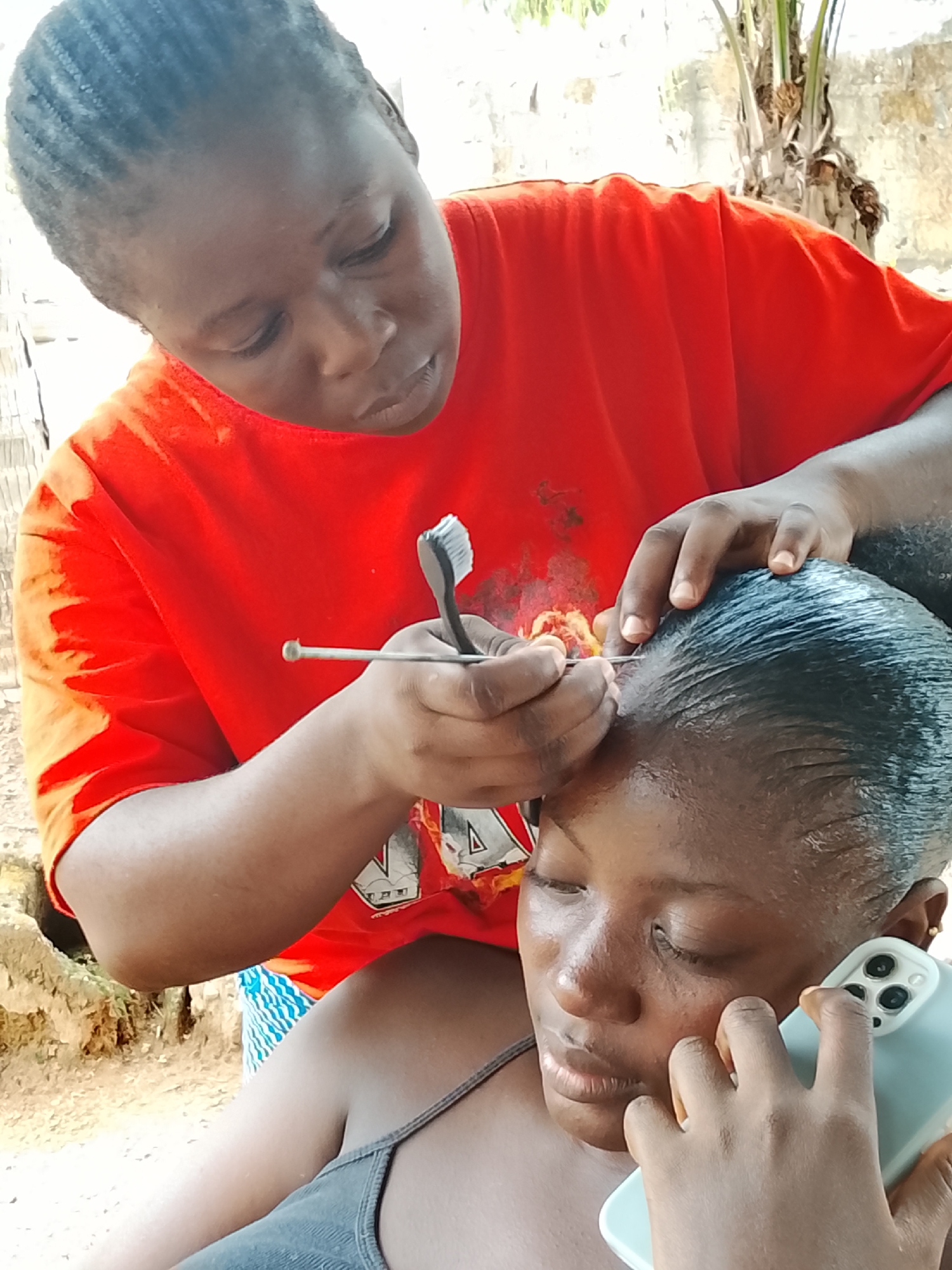 In Akwanga, Nigeria, an African woman in a red t-shirt is braiding the hair of another African woman wearing a grey tank top. The woman being braided is simultaneously on a smartphone call. The hair stylist is using a metal tool and a hair brush. The scene takes place outdoors, with tree foliage visible in the background.