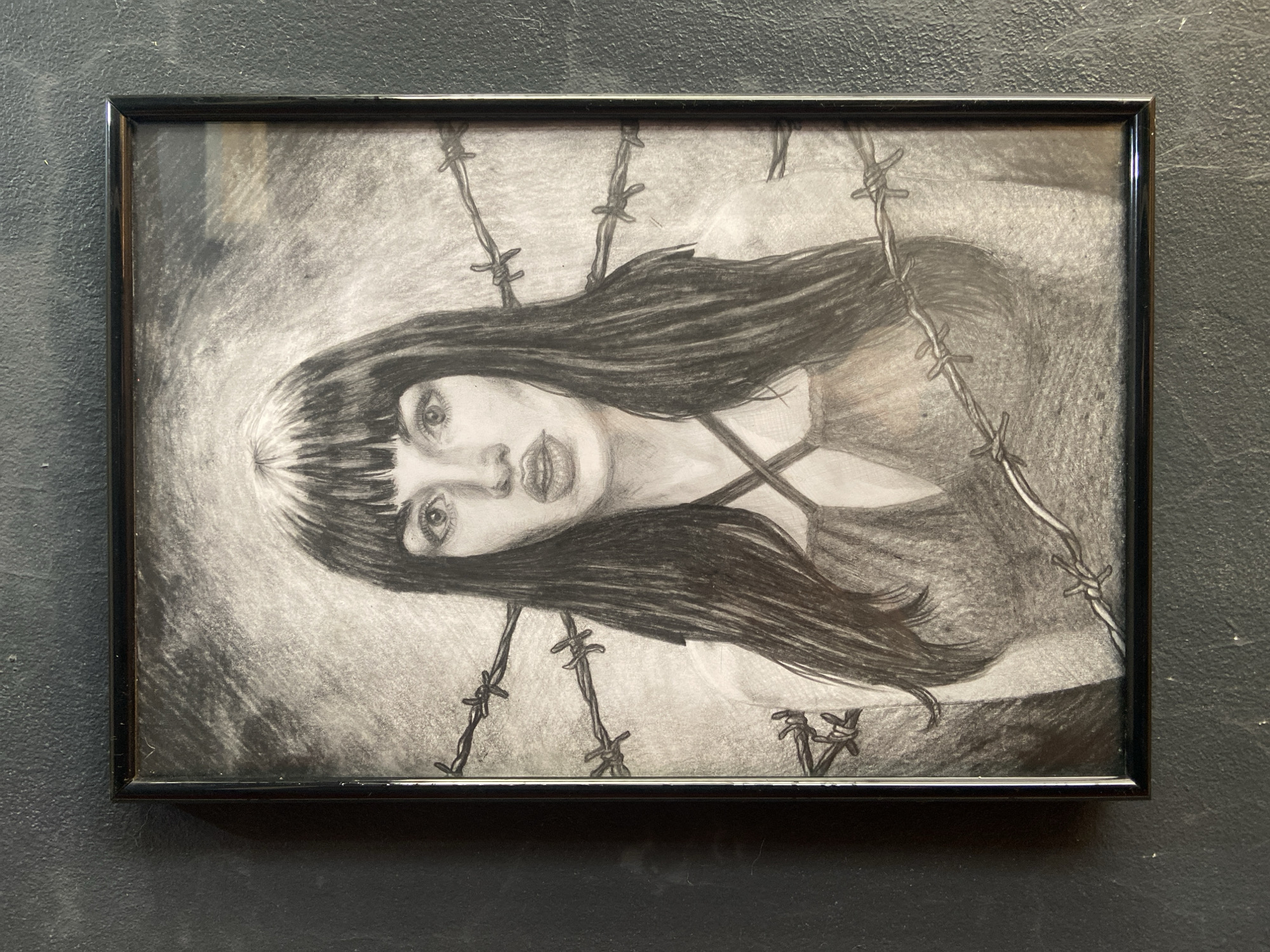 A framed charcoal or graphite drawing, oriented horizontally, depicts the head and upper torso of a young woman rendered in monochrome. The woman has long, dark, straight hair with bangs, framing a face with wide, striking eyes and full lips. Her gaze is directed slightly upward and towards the viewer. She appears to be wearing a top with thin straps.
Prominently featured throughout the drawing, intertwining with her hair and appearing over her body, are multiple strands of barbed wire, creating a sense of confinement or a barrier. The background of the artwork is a textured, shaded expanse with a lighter, almost glowing effect around the woman's head, contrasting with the darker rendering of the barbed wire and her hair. The art piece is enclosed in a simple black frame.
The drawing is displayed against a dark, textured wall, possibly concrete or stucco, suggesting an indoor setting in Buenos Aires, Argentina. No discernible activity is taking place other than the display of the artwork. The woman's expression conveys an intense or contemplative emotion. There is no visible text within the artwork or on the frame.