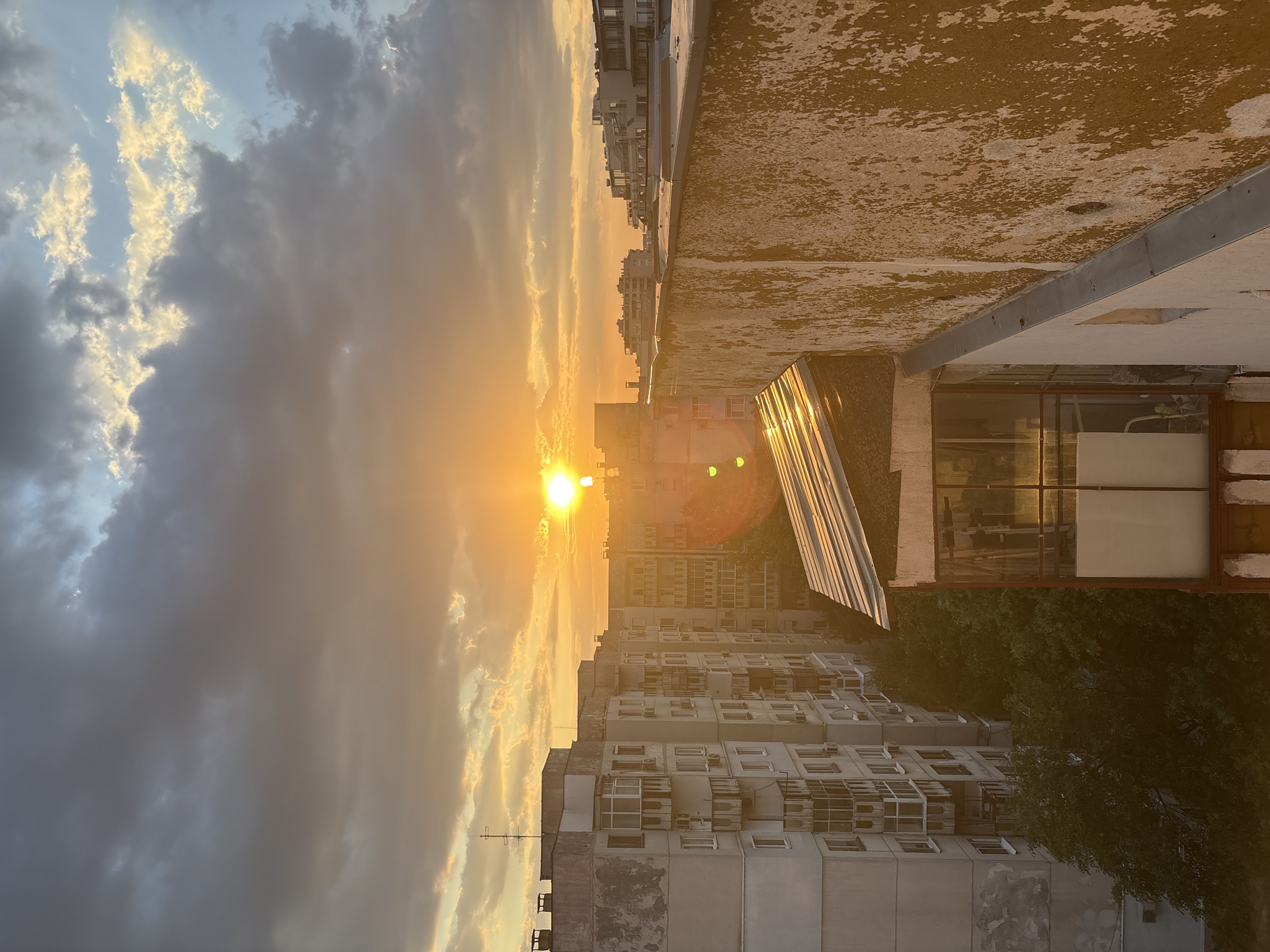 This media file captures a sunset over a residential area in Plovdiv, Bulgaria. The sun, a bright orb of light, is positioned in the lower left quadrant of the image, casting a warm, golden glow across the sky and reflecting off the surfaces of buildings. The sky is filled with a mix of dark, ominous clouds and patches of illuminated sky, suggesting a dynamic weather pattern at dusk.
The scene is dominated by the presence of tall apartment buildings, their facades a mix of concrete and visible air conditioning units, indicative of a typical urban residential setting. The buildings are densely packed, creating a layered effect that recedes into the distance. In the foreground, the rough, textured surface of a building's rooftop is visible, showing signs of wear and tear. A sloped, reflective surface, possibly a glass structure or a skylight, is also prominent on the rooftop, catching the sunlight and adding a glinting element to the scene.
The time of day is clearly sunset, evidenced by the low angle of the sun and the warm, diffused light. The weather appears to be partly cloudy, with dramatic contrast between the shadowed and sunlit portions of the sky. The overall mood is somewhat serene, with the setting sun providing a beautiful, albeit somewhat muted, display of natural light. There are no visible people or text in the image. The dense urban architecture and the specific quality of the light are the defining features of this Plovdiv scene.