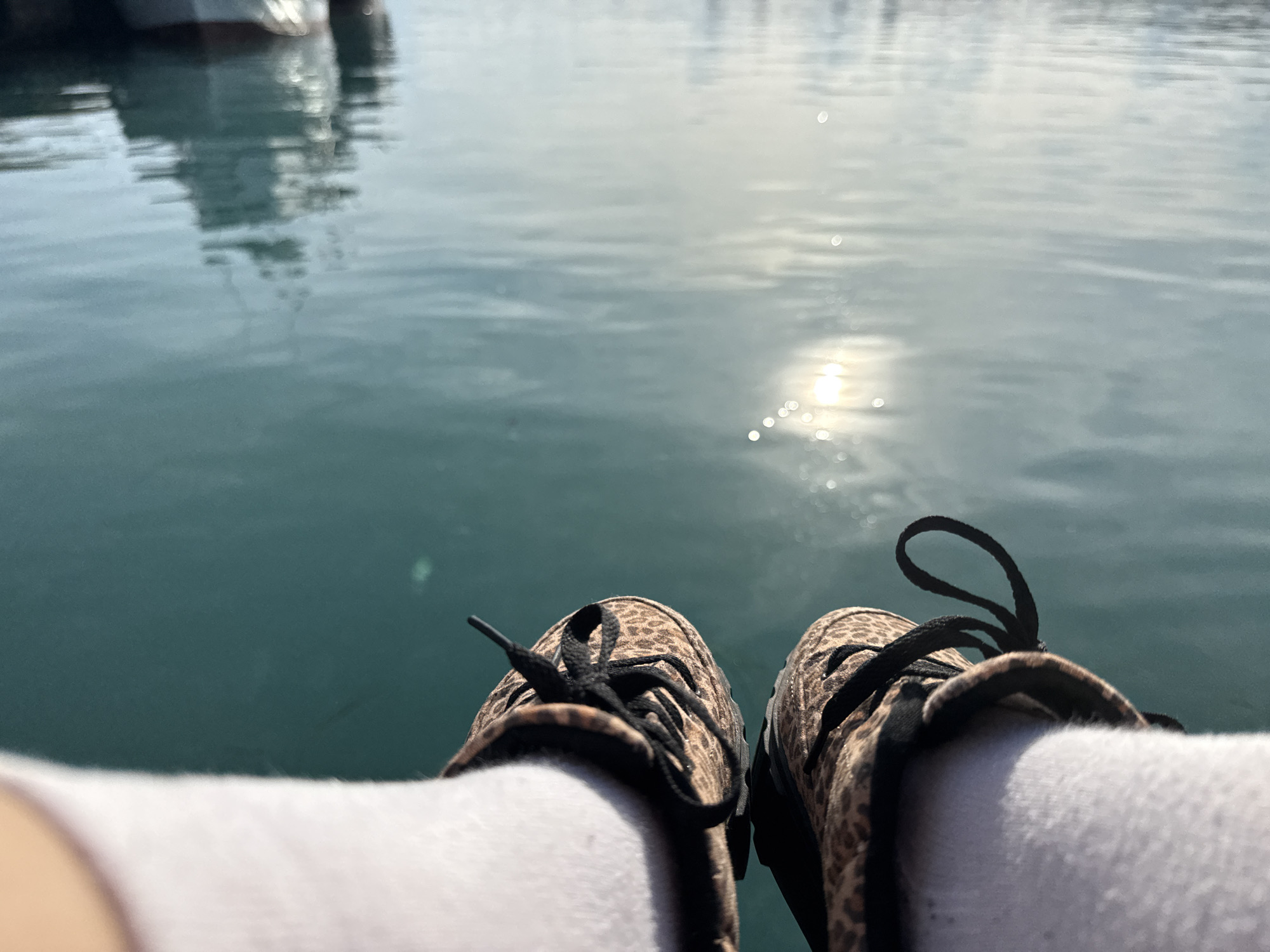 The image presents a first-person perspective of someone sitting near a body of water. The foreground features two feet wearing white socks and leopard print sneakers with untied black laces. The background reveals a body of water, possibly the Black Sea, with the sun's reflection causing shimmering light. In the distance, there is an out-of-focus building which might be located in Nesebar, Bulgaria. There are no observable people or interactions beyond the image's perspective.