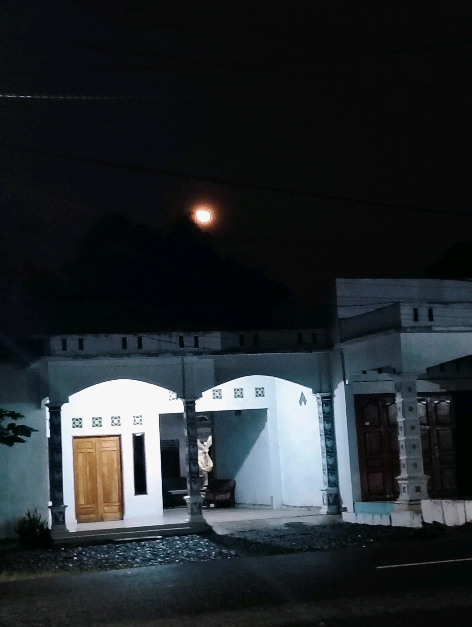 The image depicts a nighttime scene in the city of Perdamaian, Indonesia, showing a residential building under a dark sky with a bright moon. The house is a single-story structure with a white facade and a prominent arched entrance. To the left of the entrance, there are double wooden doors. Next to the doors, a narrow vertical window is visible. The building features decorative square patterns above the entrance and around the windows. The foreground shows a dark, paved surface, possibly a street or driveway, with scattered dark stones. A dark silhouette of trees or foliage is present on the left side of the image. In the background, the sky is dark, with the moon appearing as a luminous, slightly orange-hued circle. The overall atmosphere is tranquil and nocturnal.
