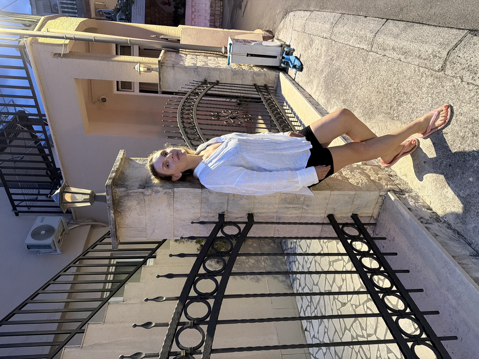 In Budva, Montenegro, a woman stands leaning against a stone pillar next to a black metal gate. She is wearing a white open-neck shirt and black shorts. Her right leg is bent and crosses over her left, and she is wearing pink flip-flops. A blue scooter is visible behind her, near a white utility box. The scene appears to be in front of a building with a staircase and an air conditioning unit visible. The lighting suggests it is either early morning or late afternoon.
