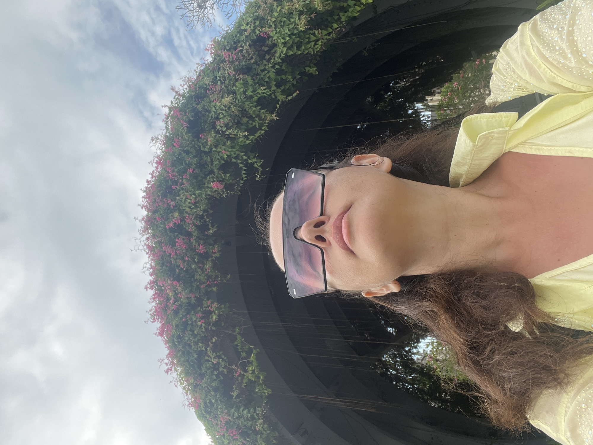An upward-angle, medium shot captures a woman wearing large, rectangular sunglasses with pink lenses. She is looking upwards, with her face partially obscured by the glasses. Her long, dark, wavy hair is visible, framing her face and falling over her shoulders. She is wearing a light yellow collared shirt with sparkling embellishments on the sleeve. The background features a dark, arch-shaped structure covered in lush green vines and small pink flowers, with hanging roots visible beneath the arch. The sky above is overcast with scattered clouds. This scene is located at the Bali Collection shopping complex in Kuta Selatan, Indonesia, specifically in the garden area with its distinctive floral archway.