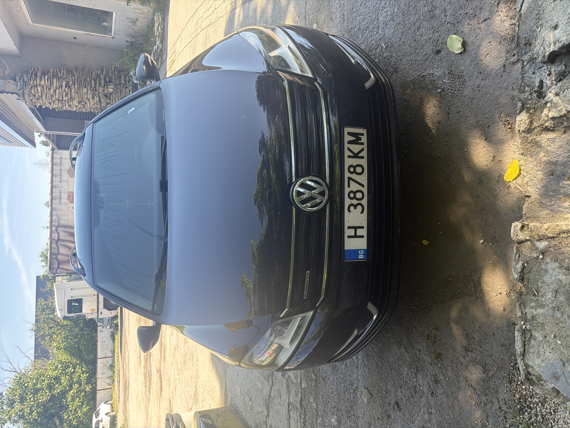 The image displays a dark-colored Volkswagen car, likely a wagon given the visible roof rails, parked outdoors on a sunny day. The car's front is prominent in the foreground, featuring the Volkswagen logo, a chrome grille, and a "BLUEMOTION" badge. The front license plate is clearly visible, displaying "H 3878 KM" with the "BG" country code and the European Union flag.
The setting appears to be a semi-urban or industrial area. To the left of the car, part of a building with a distinctive, rough-cut stone facade and a window is visible. In the background, beyond the car, there are green trees and a large white truck. A smaller white car is partially discernible behind the truck. Through the car's windshield, a sign in the distance reads "CHIMNEYS BUILDING SYSTEM".
The ground immediately around the car is a mix of concrete or rough pavement and dirt, with some scattered green and yellow leaves, and what appear to be damp or wet patches. Bright sunlight casts strong shadows, indicating it is daytime with clear weather. The overall scene suggests the car is parked in an open lot or driveway in Shumen, Bulgaria.